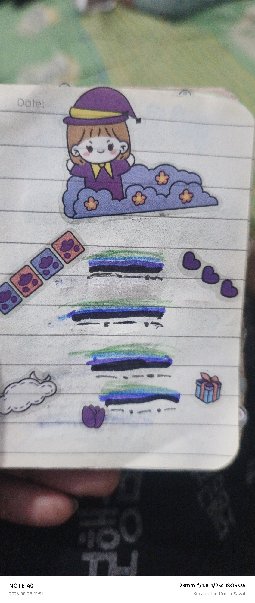 The image is a close-up shot of a notebook page with drawings and stickers. In the upper portion of the page, a cartoon drawing of a person wearing a purple hat and yellow shirt is depicted on a purple cloud with flowers. To the left of the drawing is the word "Date:" written in black ink. Below this, there are horizontal lines of colored marker streaks in blue, purple, and black, interspersed with short horizontal dashes. To the left of these streaks is a strip of square stickers featuring a pink square with a purple cloud and a purple square with a pink cloud. To the right of the marker streaks, there are four purple heart stickers arranged in a downward diagonal. Further down the page, on the left, is a sticker of a cloud with a crescent moon. Below the cloud sticker, there is a drawing of a purple tulip. To the right of the tulip is a sticker of a blue gift box with a pink ribbon. The notebook page is lined. At the bottom of the image, text indicates "NOTE 40", a date "2026.08.28 11:31", camera settings "23mm f/1.8 1/25s ISO5335", and a location "Kecamatan Duren Sawit", which is a subdistrict in East Jakarta, Indonesia. The background of the image is a blurry green and brown textured surface, likely a table or fabric.
