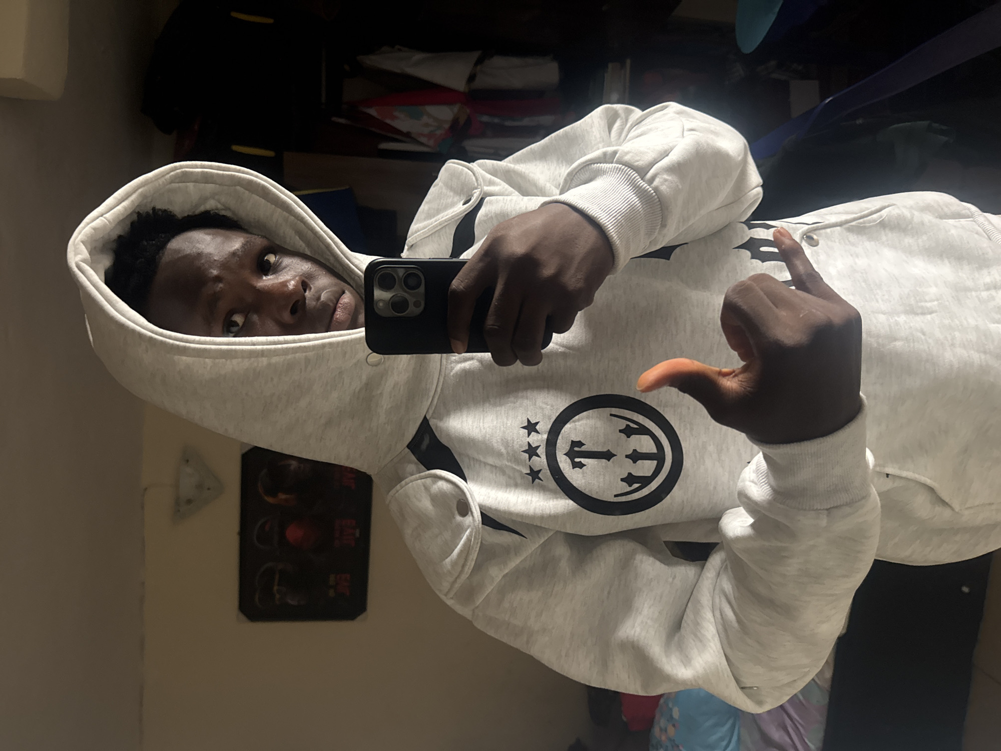 A dark-skinned young man is taking a mirror selfie in a room. He is wearing a light grey hooded sweatshirt with a black graphic logo on the chest. The logo consists of three stars above a circle with a stylized "T" in the center. He is holding a black smartphone in his right hand, angled to capture his reflection. His left hand is extended towards the camera, with his thumb up and his index and pinky fingers extended in a gesture. The background of the room is dimly lit, with a closet filled with clothing visible on the right. A poster with three figures is on the wall to the left. The image appears to be taken in a residential setting, possibly a bedroom, within the city of Ihiagwa, Nigeria.