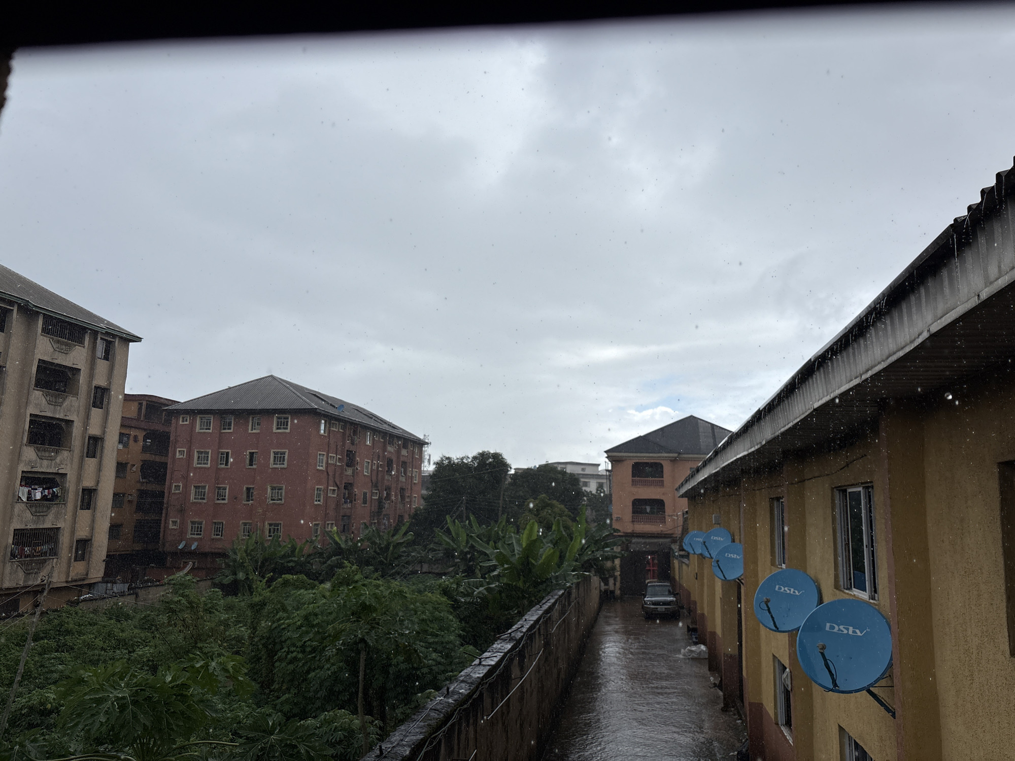 The image depicts a street scene in Onitsha, Nigeria, during a rainfall. The sky is overcast and gray, with visible raindrops falling. Several multi-story buildings are visible, including a light brown apartment building on the left with balconies and laundry hanging out, and a reddish-brown building behind it with numerous windows. On the right, a yellow building with a dark roof runs along the street, featuring several blue satellite dishes mounted on its exterior. A narrow, wet asphalt street runs between the buildings, with a parked car visible in the distance. Lush green foliage, including banana trees, lines the street and the area between the buildings, suggesting a tropical environment. The scene is captured from a slightly elevated perspective, looking down onto the street and surrounding structures.