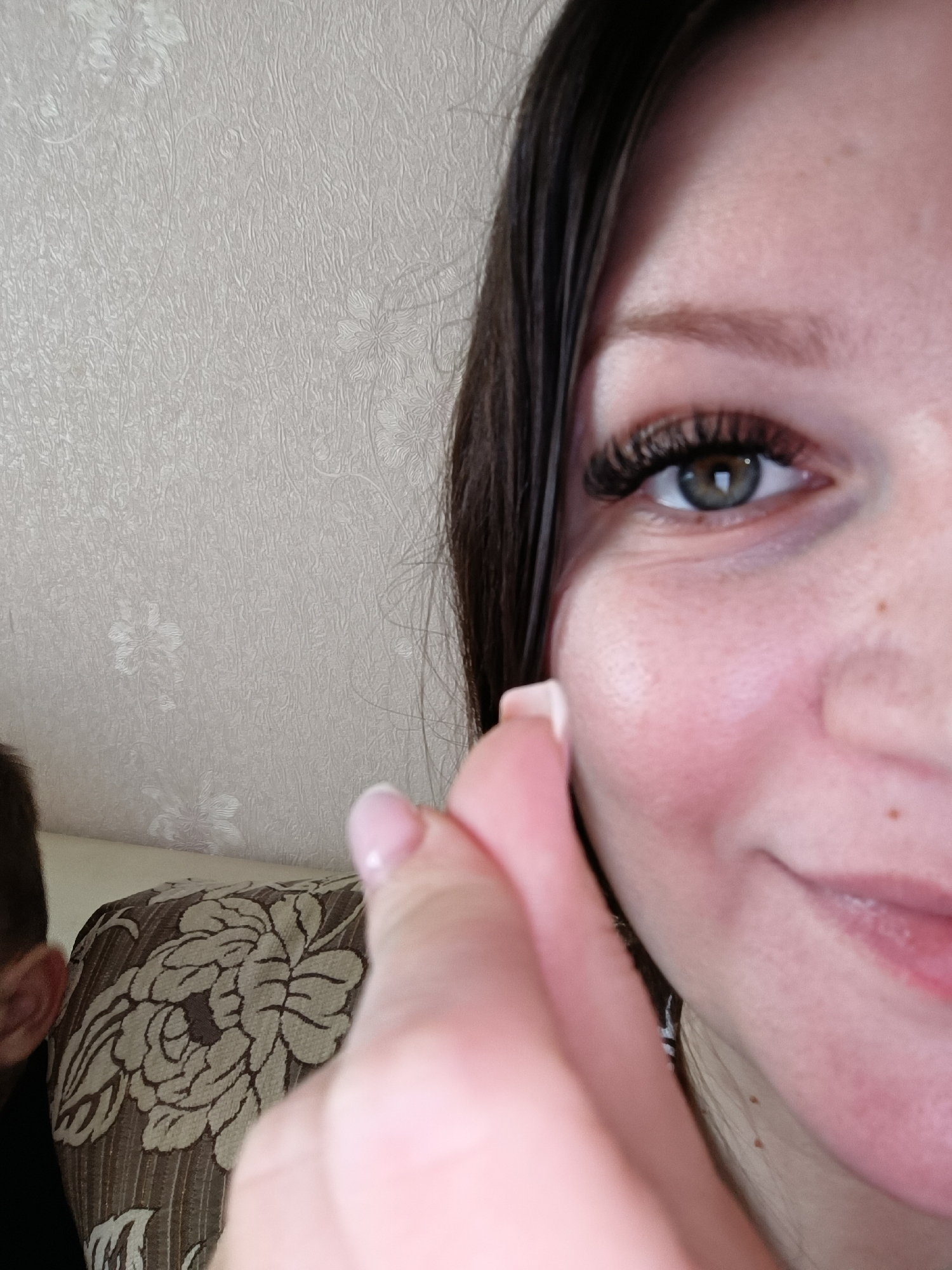 A close-up image shows a portion of a person's face and hand. The person has brown hair, a light complexion, and hazel eyes with long, dark eyelashes. The skin shows freckles. The person's right hand is raised toward their face, with manicured nails that have a light pink polish. In the background, part of a patterned upholstered chair is visible, along with a textured off-white wall. There is a second person partially visible to the left, only their ear and the back of their head are visible. There is no information in the image to indicate the location is Tobolsk, Russia.