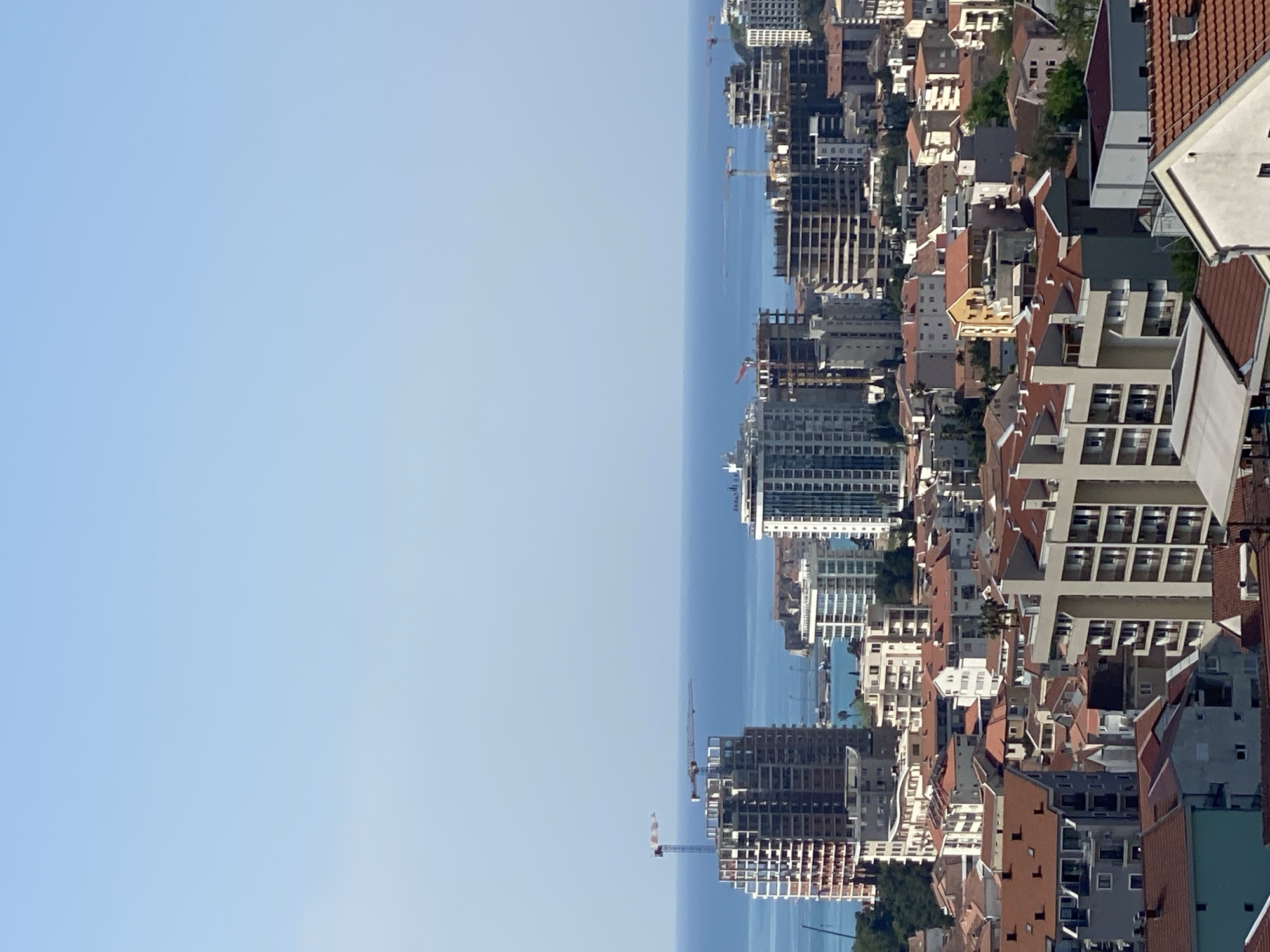 The image displays a panoramic view of a coastal city, identified as Budva, Montenegro, under a clear, bright blue sky. The scene is heavily populated with urban architecture, showcasing a mix of completed structures and numerous buildings under active construction.
In the foreground and midground, a dense array of buildings stretches across the landscape, descending towards the coastline. These include multi-story residential and commercial buildings, many featuring red or orange terracotta roofs typical of the Mediterranean region. Interspersed among the established buildings are several large construction sites, with visible concrete frameworks, scaffolding, and bare rebar, indicating extensive urban development. Multiple tall construction cranes are prominently featured across the cityscape, emphasizing the ongoing building boom.
The deep blue sea forms a distinct horizon line, separating the land from the clear sky. A few small boats or ships can be seen in the distant waters. The sky itself is a vibrant, cloudless blue, suggesting a sunny day, likely late morning or early afternoon given the bright, even lighting on the buildings. There are no people visible in the image, nor is there any discernible text. The overall impression is one of a rapidly developing coastal metropolis.