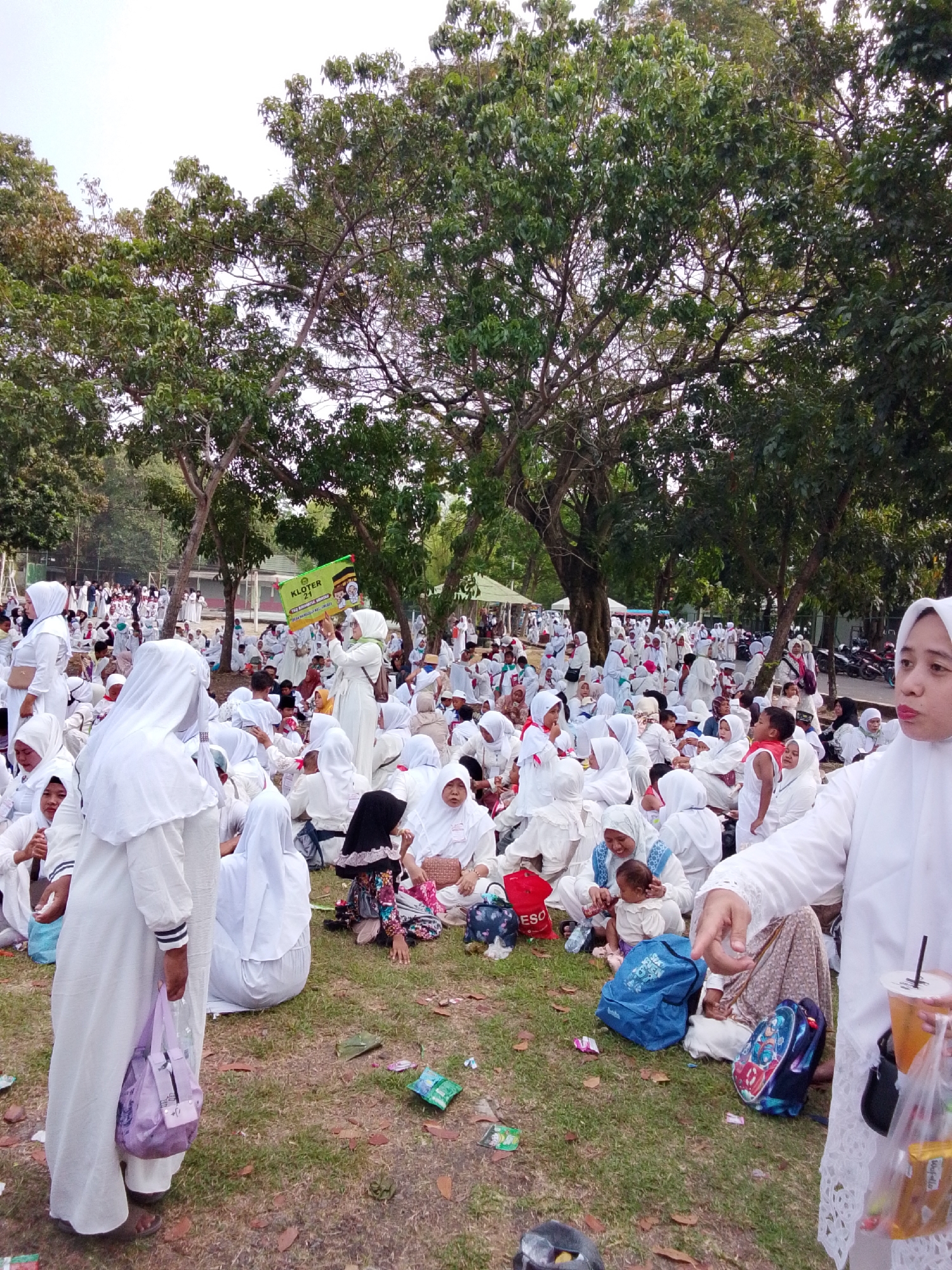 In Cirebon, Indonesia, a crowd of people, many wearing white Islamic attire, are gathered on a grassy area under the shade of trees. The scene appears to be an outdoor event or gathering. In the foreground, a woman in a white hijab and dress is holding a clear plastic cup filled with an orange beverage and a plastic bag with snacks. Several other individuals are seated on the ground, some in groups, engaged in conversation or resting.
Further back, a large congregation of people in white is visible, suggesting a religious or cultural event. Some individuals are standing, while others are seated on the grass. A sign with green and yellow lettering, reading "KLOTER 21" and featuring an image of a man in a white robe, is being held up by a woman in the middle ground. In the background, more trees and some buildings are visible, along with parked motorcycles on a paved area to the right. The overall atmosphere appears peaceful and communal, with a mix of adults and children present. The ground is scattered with leaves and some pieces of trash, indicating an outdoor setting that may have seen recent activity.