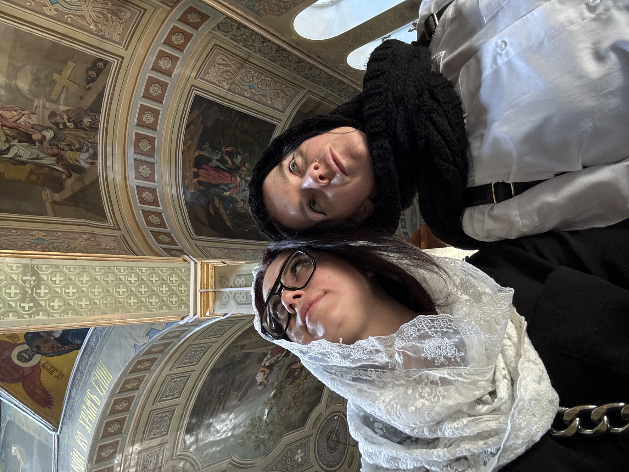 Two women are looking up at the ceiling of a church. The woman on the left wears a white lace headscarf and black clothing, with large black-rimmed glasses. The woman on the right wears a black headscarf and a white shirt. Both women have their heads tilted upwards, gazing at the ornate painted ceiling and architectural details of the church. The ceiling features religious paintings and decorative elements. The lighting suggests an interior setting, possibly with natural light filtering in from windows.