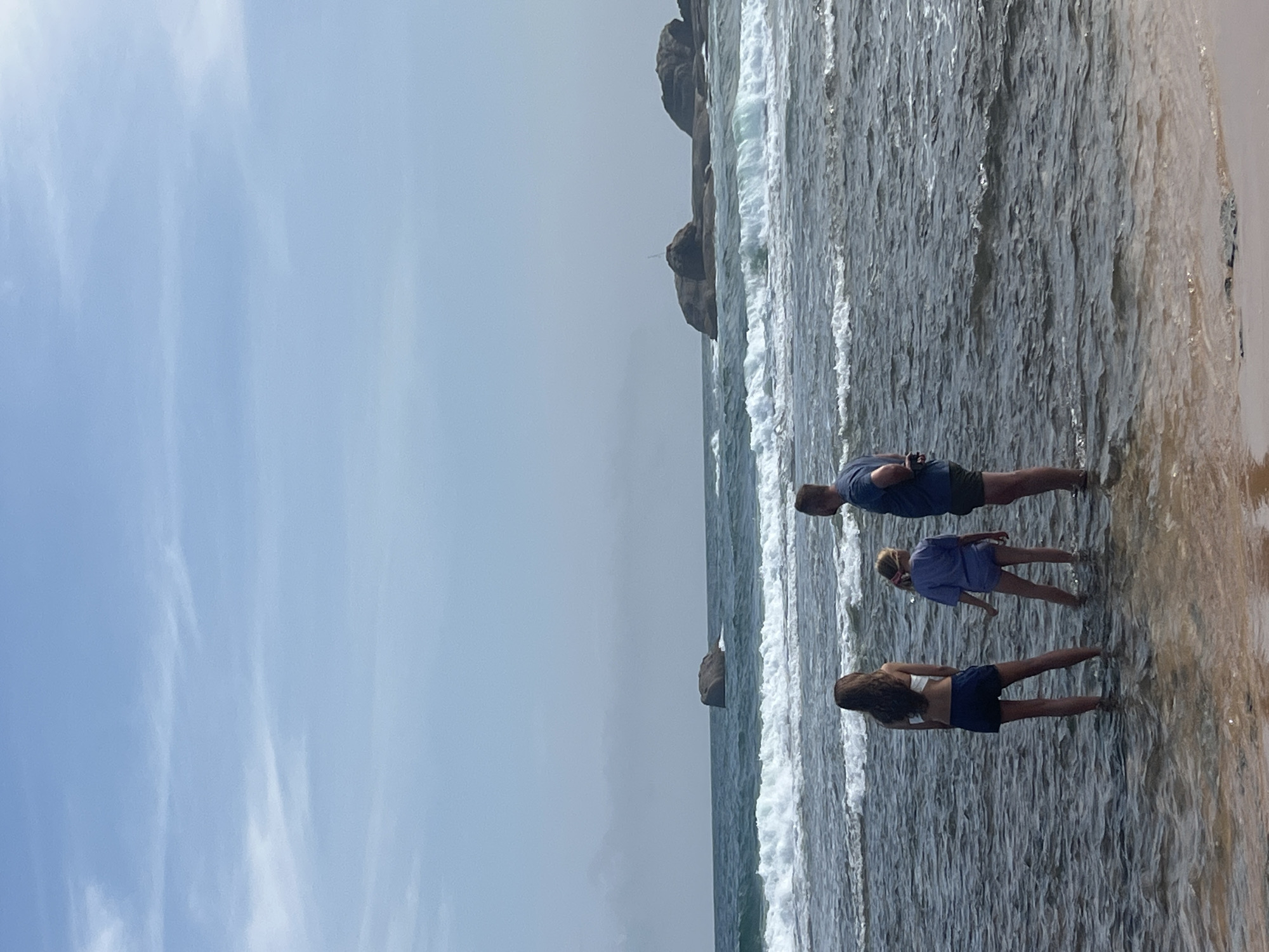 In a scene at the coast of Hikkaduwa, Sri Lanka, three individuals are standing in the shallow water. The image captures a moment of leisure, with the sea gently lapping at their feet. The water is a mix of grey and blue, with white foam from the waves indicating moderate activity.
The subjects appear to be two adults and one child, their bodies oriented towards the sea, suggesting they are facing the ocean or engaged in an activity within it. One person is wearing a blue t-shirt and dark shorts, another a purple t-shirt and dark shorts. The third person is wearing a white bikini top and dark shorts. Their postures suggest a relaxed atmosphere.
In the background, large rock formations rise from the water, characteristic of coastal landscapes. The sky is a pale blue with wispy white clouds, indicating daytime, likely late morning or early afternoon due to the brightness. The overall mood appears calm and serene, with a focus on the natural beauty of the beach and the interaction of the people with the ocean. No visible text is present in the image.