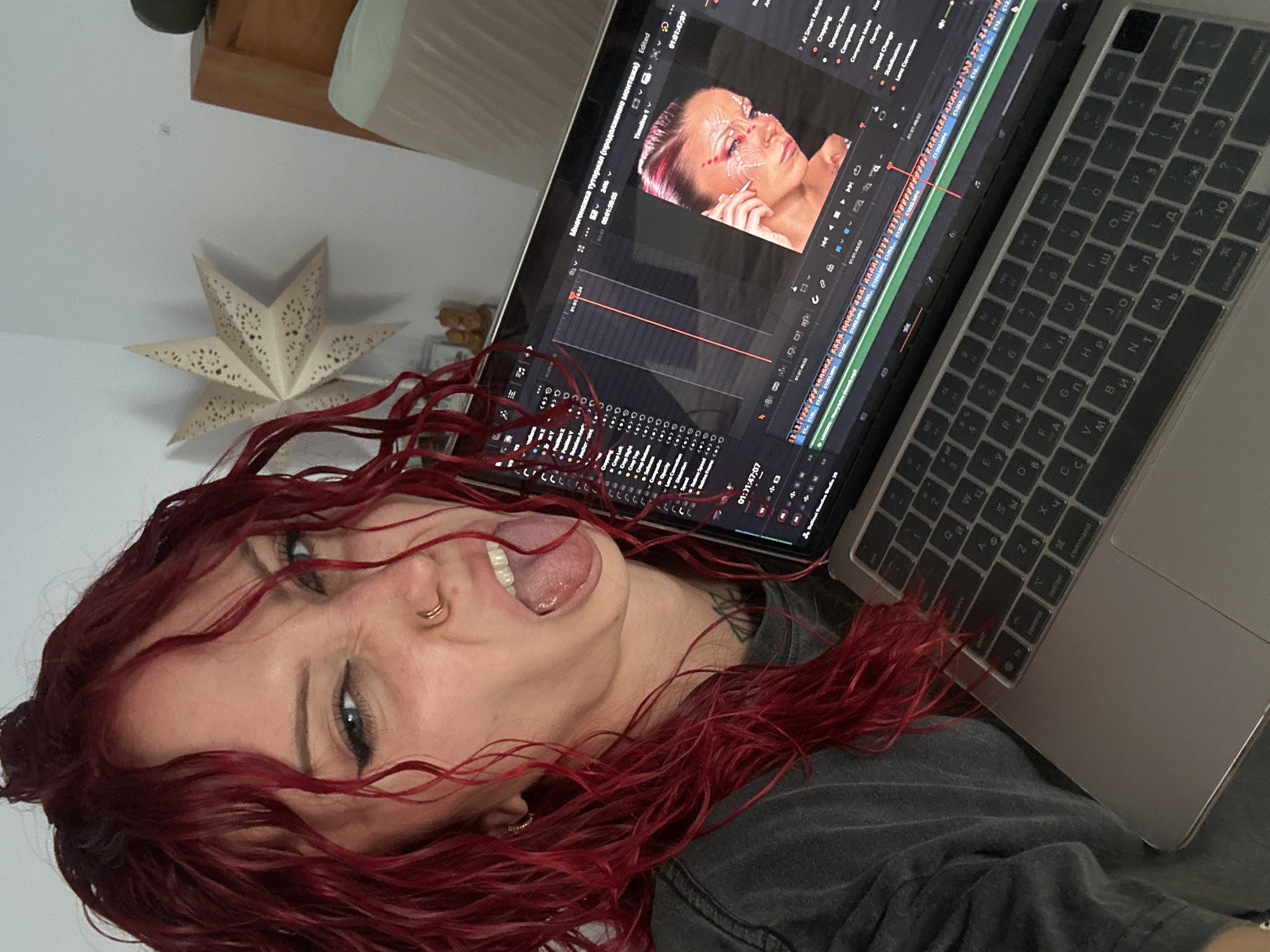 A person with red hair is positioned in front of an open laptop, with the screen displaying video editing software featuring an image of a person. The laptop is at an angle relative to the camera. The person is sticking out their tongue and making a playful face. A star-shaped decoration is visible in the background. The keyboard of the laptop is visible and appears to have Cyrillic characters. There is no information to suggest a specific location or connection to North Kuta, Indonesia.
