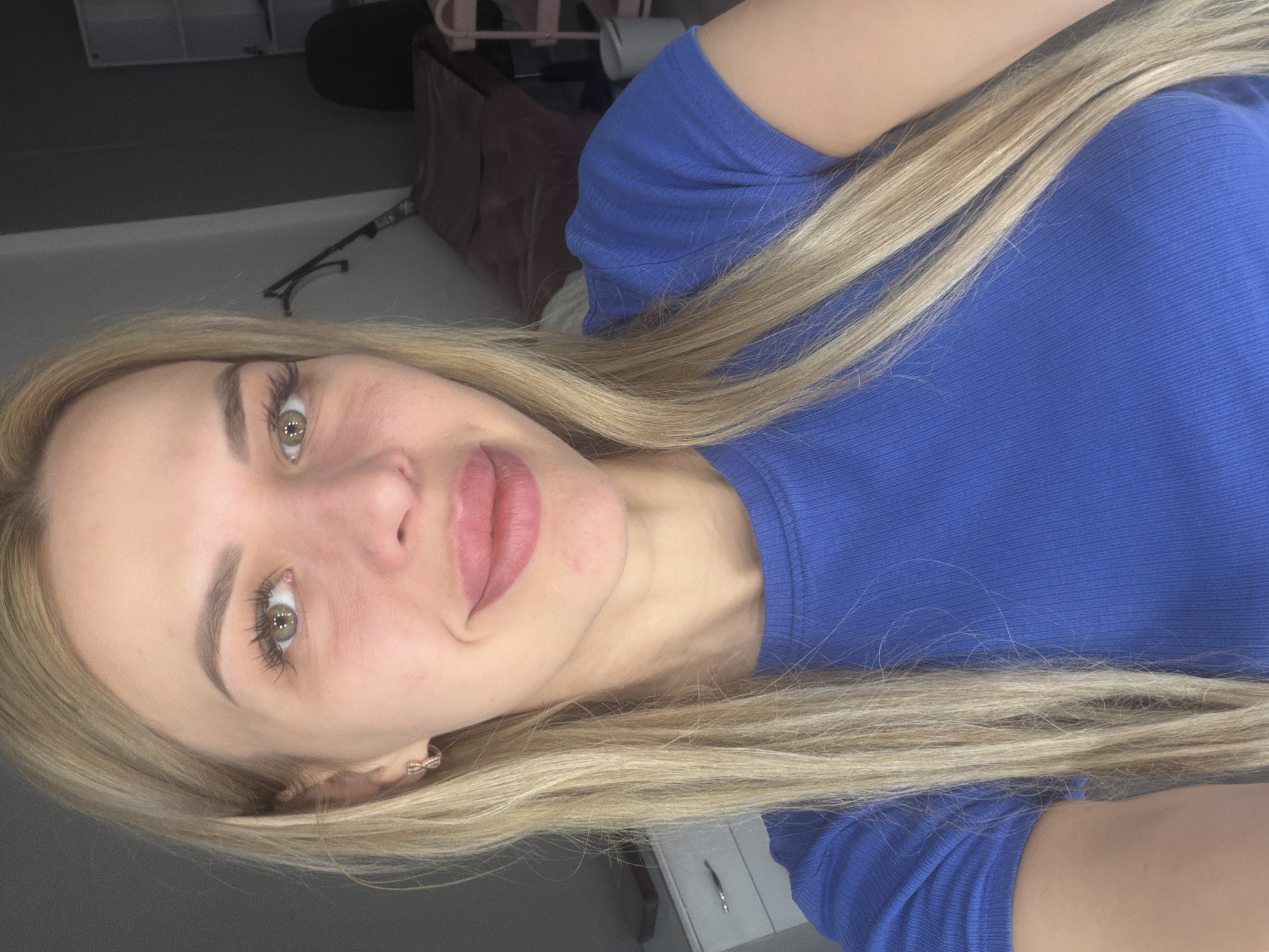 A close-up selfie shows a fair-skinned blonde woman with light green eyes and full lips, wearing a royal blue ribbed t-shirt. Her hair is straight and falls around her shoulders. She is smiling slightly, with a faint blush on her cheeks. The background is dimly lit and appears to be an indoor setting, possibly a room with furniture and shelves visible in the distance. The image does not provide any specific information to determine its location within the city of Kyiv, Ukraine.