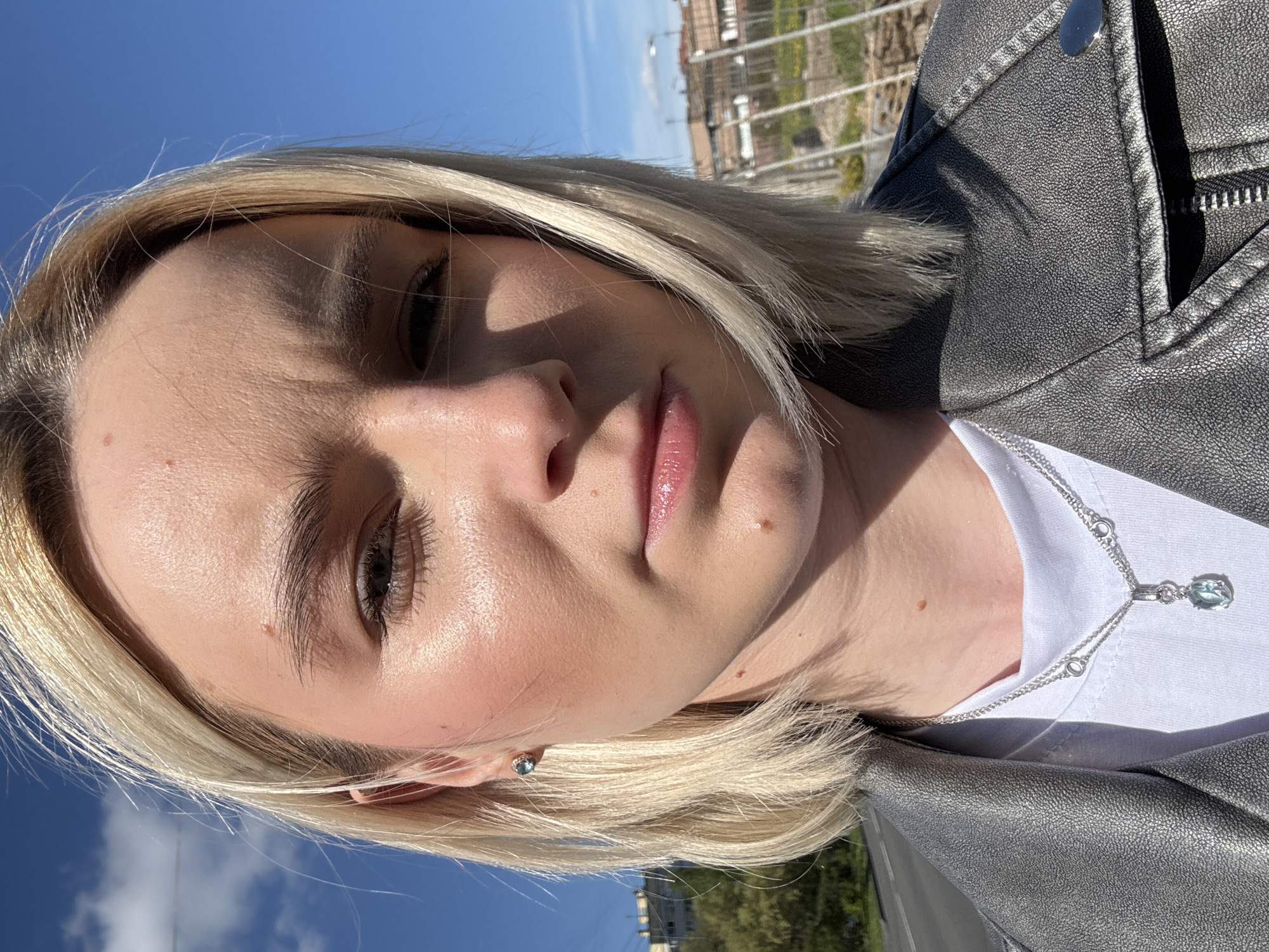 Here's a description of the image:
A close-up, angled shot of a woman with blonde, shoulder-length hair. She is wearing a silver-colored leather jacket over a white t-shirt and a silver necklace with a light blue pendant. The background shows a bright blue sky, what appears to be a fence or railing, a building, and greenery, likely trees, suggesting the image was taken outdoors on a sunny day. Based on the architectural style, the building may be in the city of Saint Petersburg, Russia. The focus is on the woman's face, capturing her features and complexion.