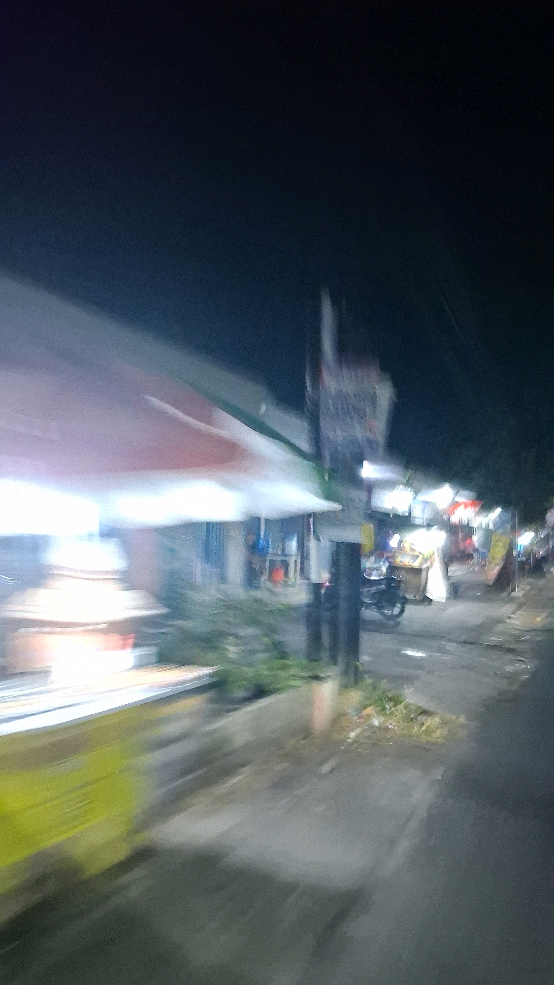 The image depicts a street scene at night in Jogotirto, Indonesia, characterized by motion blur, suggesting the photograph was taken from a moving vehicle. On the left, a brightly lit food stall or cart with a yellow base and a white and red awning is visible. Adjacent to it, further down the street, are more stalls or shopfronts with various lights and offerings. A motorcycle is parked in the middle ground. The road surface appears wet, reflecting the ambient light. The background is dark with some blurred greenery and indistinct structures. The overall impression is of a busy, illuminated street at night, possibly a market or a commercial area.