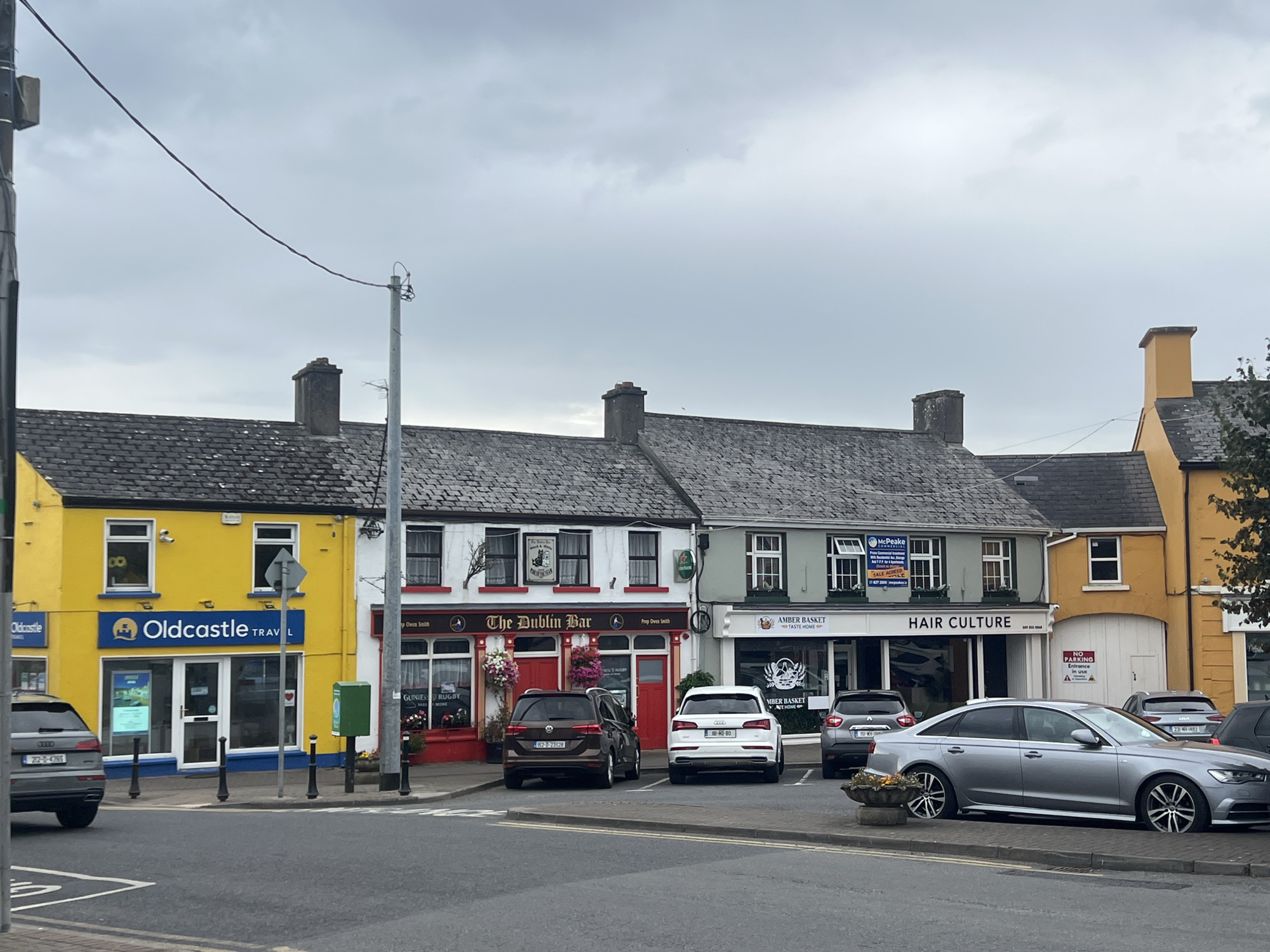 The image depicts a street scene in a European town, likely Ireland, given the license plates and signage. The sky is overcast. In the foreground, a grey sedan is parked alongside a curb, with a planter to its left. Behind it, several other cars are parked in a line. The street itself has white directional lines.
The middle ground features a row of businesses with distinct facades. From left to right, the first is a bright yellow building with "Oldcastle Travel" signage. Next to it is a white building with a red facade, identified as "The Dublin Bar." Further to the right is a light green building with "Amber Basket" and "Hair Culture" signage. The last visible building on the right is yellow, with white garage doors. There are multiple windows and doorways visible on these buildings, some with decorative elements like flowers. Several cars are parked in front of these establishments.
The background shows the roofs of these buildings, characterized by grey slates and chimneys. Electrical poles and wires are also visible. The overall impression is a commercial street on a cloudy day.