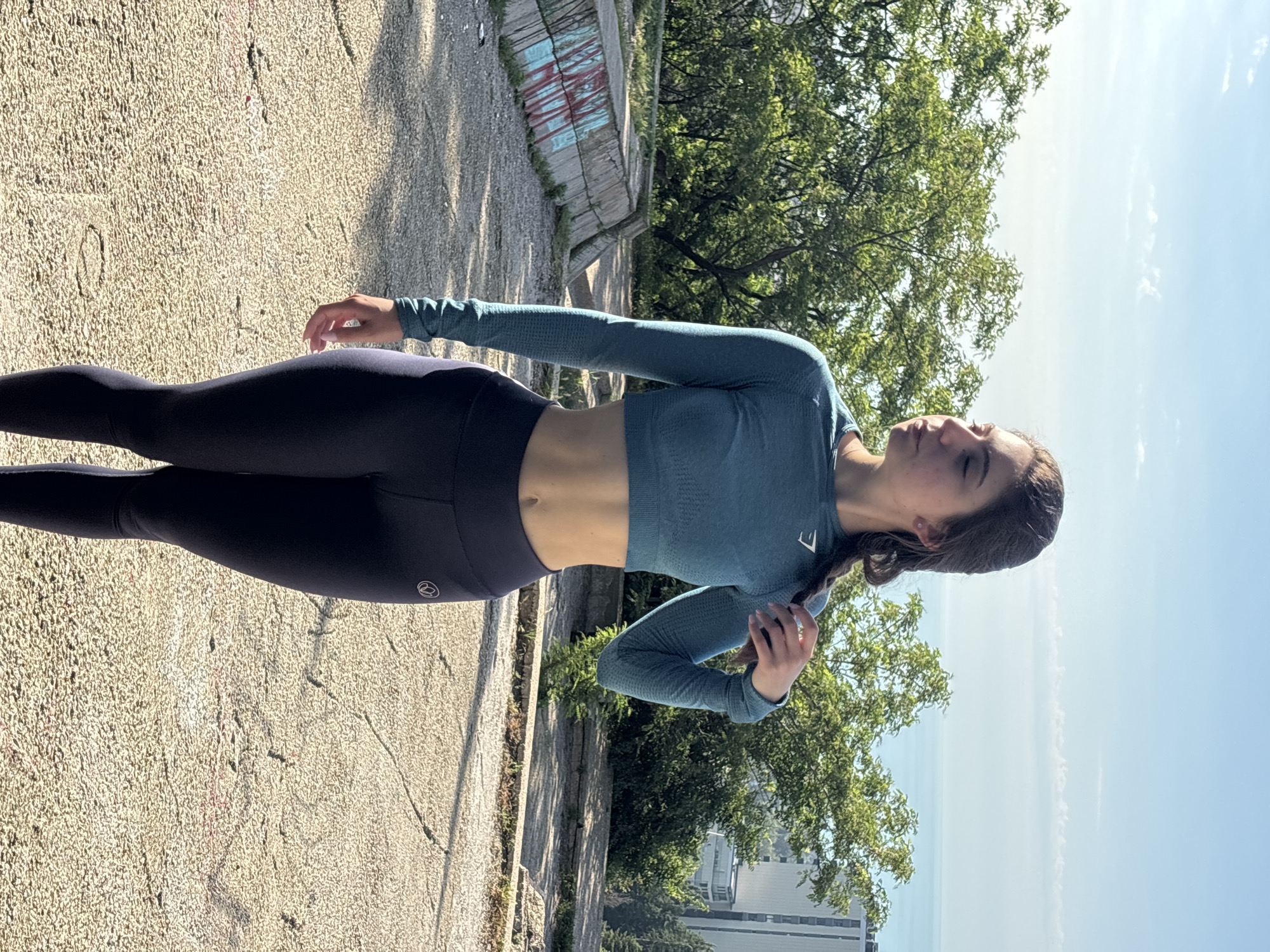 This full-body outdoor portrait features a young woman standing on a textured, light-colored path. She is dressed in athletic wear, including a long-sleeved, cropped teal-blue top and dark blue leggings. Her dark hair is pulled back, and her head is tilted upwards with her eyes closed, conveying a sense of peace or enjoyment of the sunlight.
The setting appears to be a park or promenade overlooking a body of water in Varna, Bulgaria. A low concrete wall with faded red and blue graffiti runs parallel to the path on the left. Behind this, lush green trees dominate the midground. In the distance, a calm body of water, likely the Black Sea, stretches under a bright blue sky dotted with scattered thin white clouds. Several buildings are visible on the distant horizon line.
The image is captured on a clear, sunny day, indicated by the strong natural light and distinct shadows. The woman's posture is relaxed, with her arms resting by her sides, one hand gently touching her hip. A small, light-colored, stylized 'E' logo is visible on the left side of her chest on the top. Another circular, light-colored logo, difficult to decipher precisely, is present on the outer thigh of her left legging. The overall atmosphere is calm and serene, suggesting a moment of relaxation outdoors.