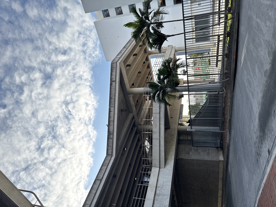 The image depicts an exterior view of the Hialeah Market Metrorail Station in Hialeah, Florida, United States of America, captured from a ground-level perspective.
The upper portion of the frame features a bright blue sky with extensive coverage of textured, patchy altocumulus or stratocumulus clouds. Sunlight indicates daytime.
The dominant subject is a multi-level concrete structure, characteristic of a transit station and parking garage. This structure exhibits exposed concrete, multiple support columns, and horizontal beams forming the undersides of elevated platforms and walkways. Metal railings line the edges of these elevated sections. Recessed lighting fixtures are visible on the concrete ceilings. A small, dark bird is perched on the top edge of the uppermost concrete slab. A sign, partially visible on a lower concrete column, reads "Hialeah Market."
To the right of the concrete structure, a modern, multi-story white building with dark-framed rectangular windows on its upper floors is visible. Several mature palm trees with slender trunks and green fronds are situated in the mid-ground, partially obscuring the white building and bordering the concrete structure.
The foreground consists of an asphalt or concrete ground surface, with a narrow strip of reddish-brown brick pavers on the far left. A dark metal fence with vertical bars runs across the mid-ground, separating the immediate foreground from the base of the concrete structure and a small grassy area. Behind this fence, a concrete staircase or ramp descends from the elevated structure.
Two individuals are partially visible behind the metal fence, near the base of the staircase/ramp, in the vicinity of the "Hialeah Market" sign. One individual appears to be wearing a dark top. Their specific actions are not discernible. A light-colored vehicle is partially visible behind the fence and concrete structure, on a lower level. Further in the background, utility poles and distant structures are faintly visible.