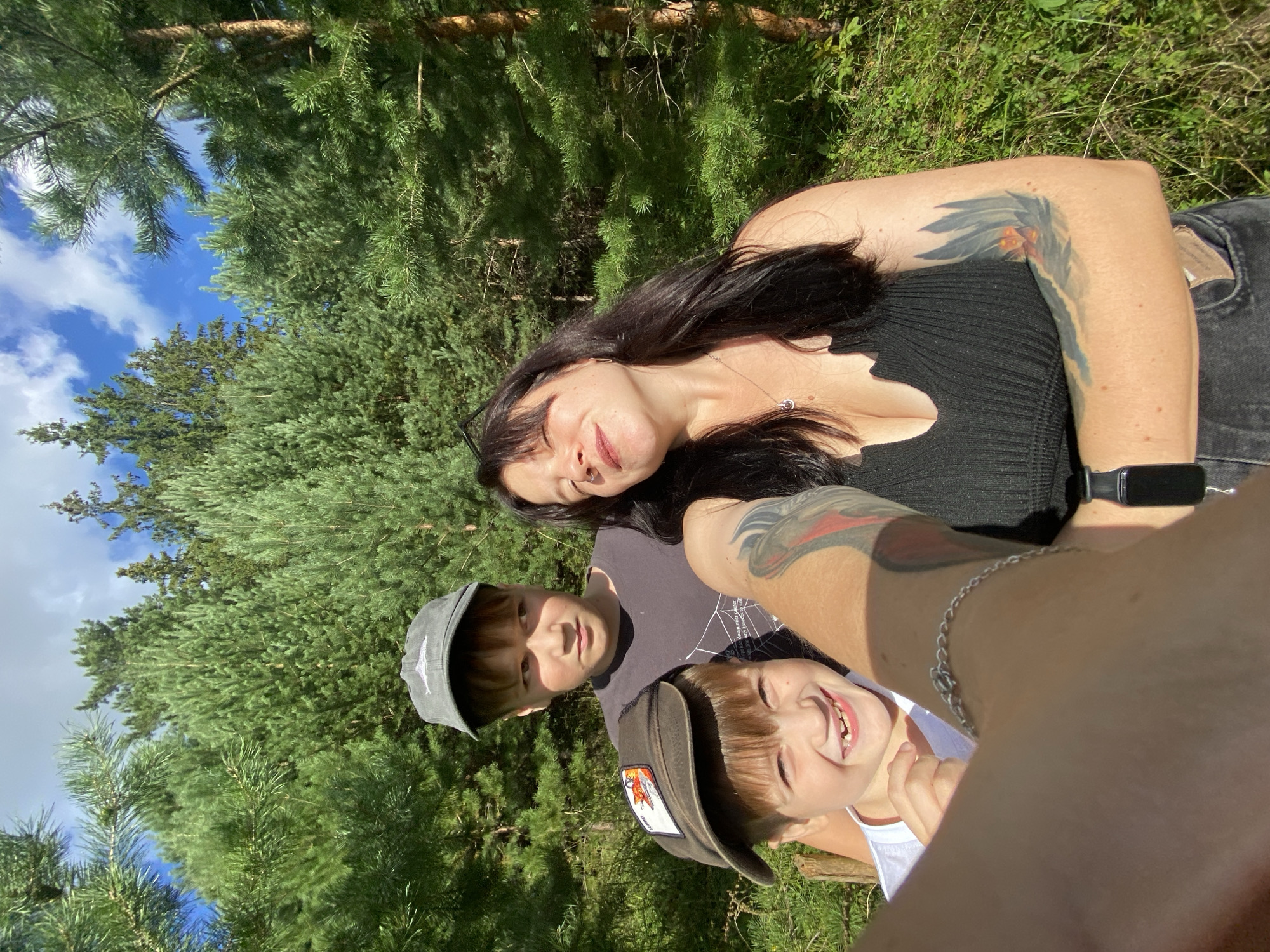 In an outdoor scene set in the city of Foky, Russia, a woman and two young boys are captured in a selfie. The woman, with long dark hair and wearing a black sleeveless top and dark jeans, is in the foreground. She has a necklace and a nose ring, and tattoos on her left arm and shoulder. Her eyes are closed, and she is smiling. To her left and slightly behind her, a boy wearing a grey baseball cap and a dark t-shirt looks to his left. Next to him and in front, a younger boy with a dark baseball cap showing a fox patch is smiling widely, displaying missing front teeth. The background consists of a dense forest of pine trees under a bright blue sky with scattered white clouds. The lighting suggests it is daytime, with sunlight illuminating the scene.