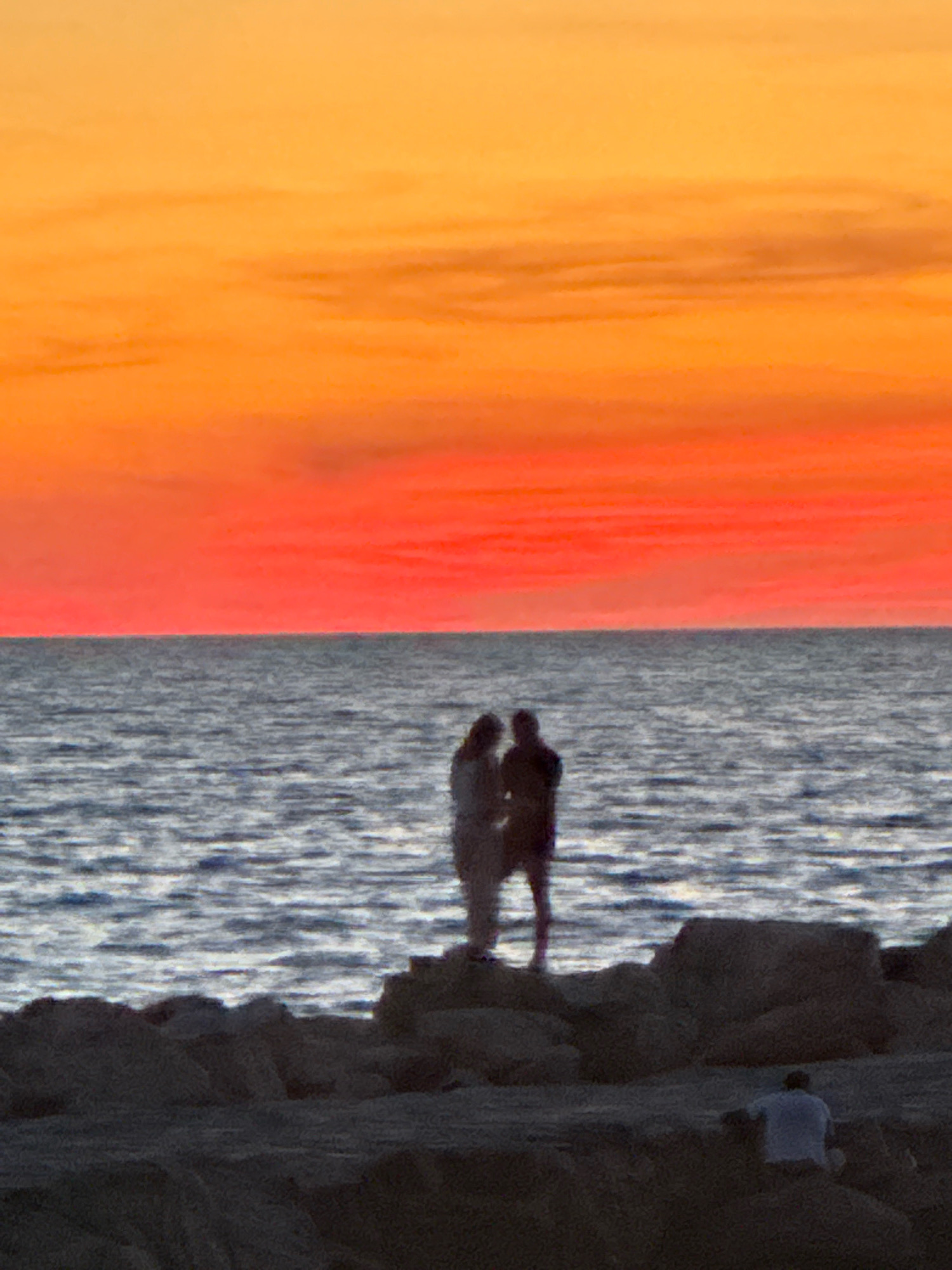 At dusk, two silhouetted figures stand close together on a rocky shoreline, gazing out at the sea. The sky above is a vibrant gradient of orange and red, indicative of a sunset. The water is calm with gentle ripples reflecting the colorful sky. In the foreground, scattered rocks form the irregular coastline. Further to the right, another silhouetted figure, wearing a white shirt, is crouched on the rocks. The scene evokes a sense of tranquility and natural beauty.