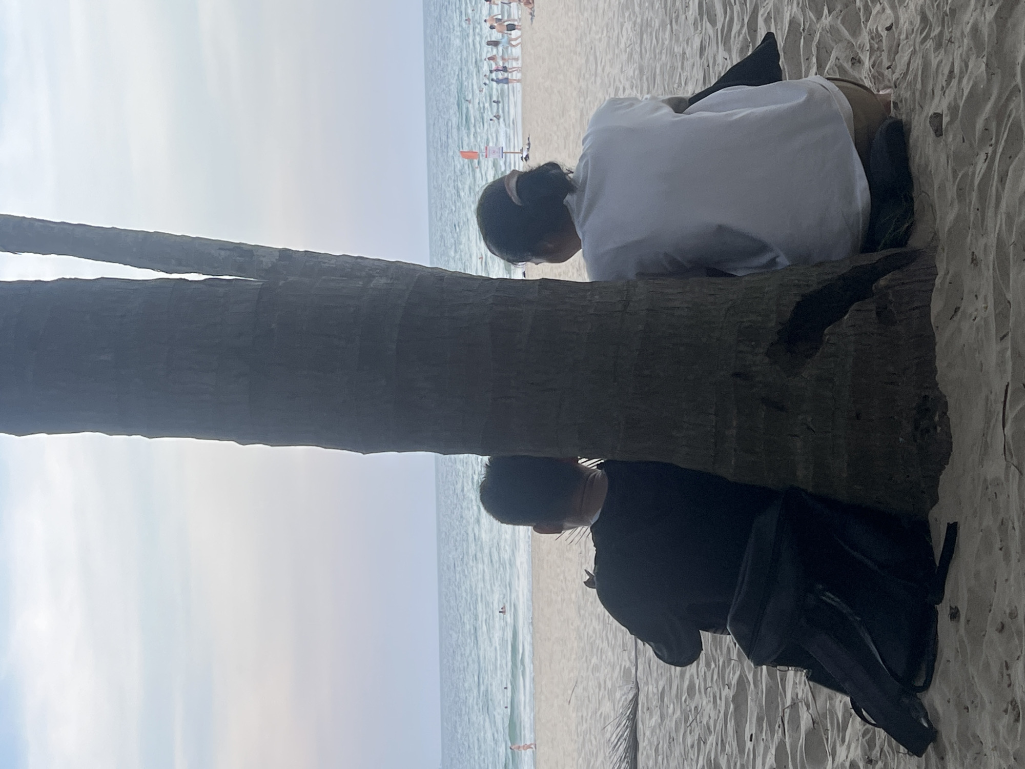 Two individuals are seated on a sandy beach in Đà Nẵng, Vietnam, leaning against a palm tree. The person on the left wears a black long-sleeved shirt and has a black backpack at their side. The person on the right wears a white t-shirt and brown shorts. They are both facing away from the camera, looking towards the ocean. The sea stretches out before them with gentle waves. In the background, other beachgoers are visible, some in the water and others on the sand, and a red flag is planted on the beach. The sky above is overcast with soft clouds, creating a muted natural light.