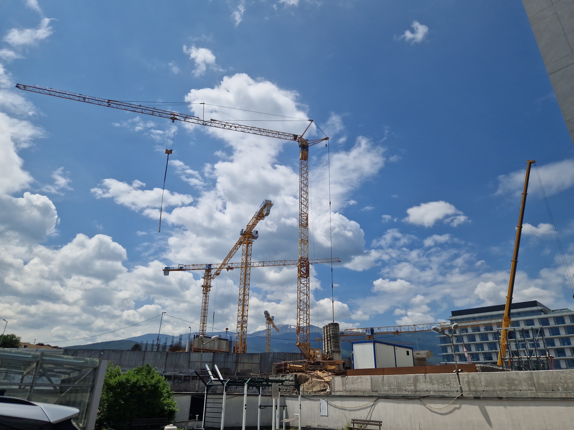 The image depicts a construction site in Sofia, Bulgaria, characterized by multiple yellow tower cranes against a backdrop of a blue sky with scattered white clouds. A building with a modern architectural design, featuring a glass facade, is visible in the background. A low mountain range is also observable on the horizon. In the foreground, there is a concrete wall and some vegetation. The overall scene suggests active urban development and construction activities.
