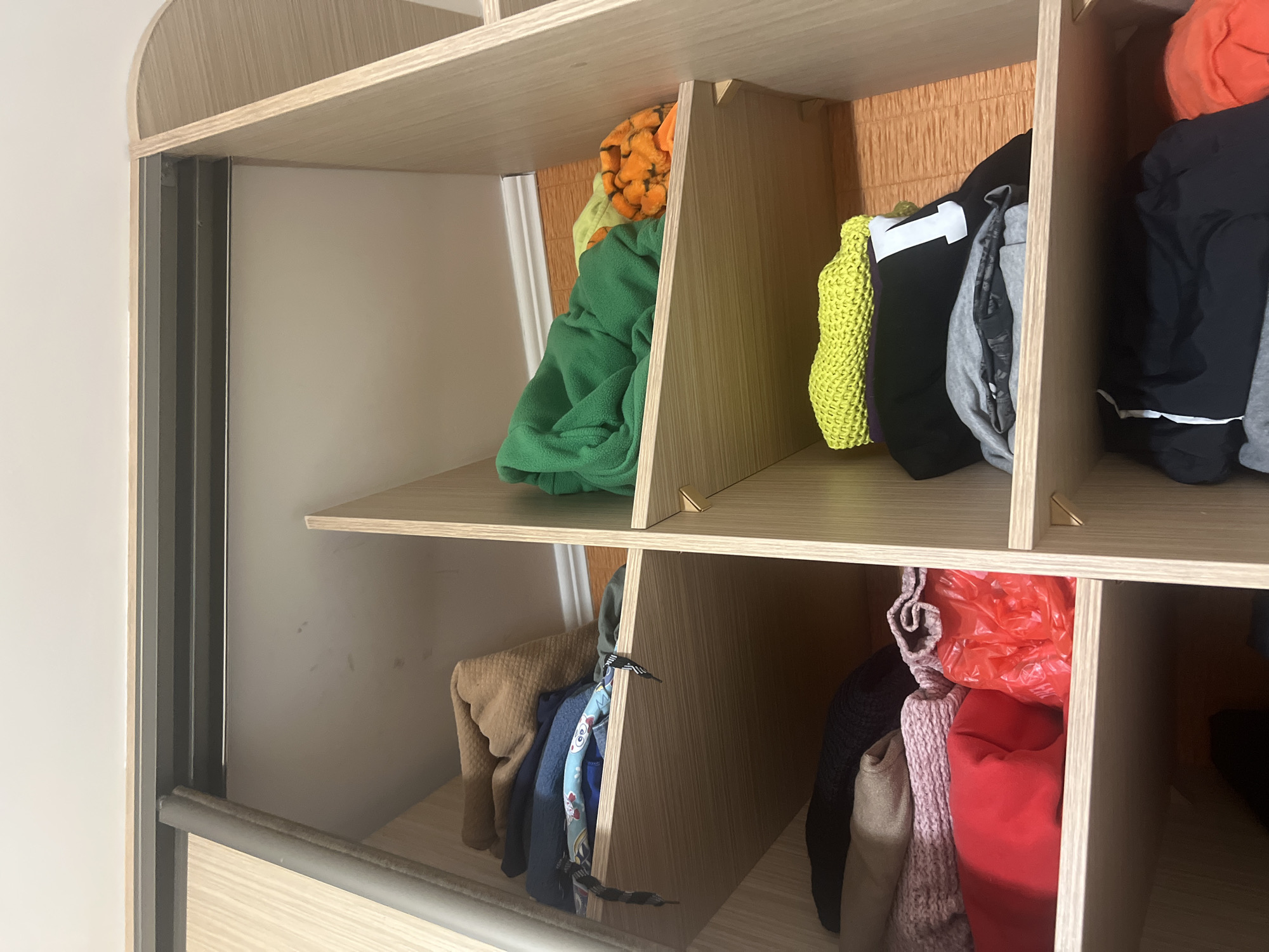 This is a low-angle, indoor shot of a closet filled with neatly folded clothing. The closet is made of light wood and has several shelves. On the top shelf of the left section, there are a few stacked items of clothing, including a brown, textured garment and a blue garment with a graphic print. The shelf below it is empty. On the bottom shelf of the left section, there are several folded garments: a dark blue or black sweater, a pink knit item, a red item, and a crinkled red plastic bag.
The right section of the closet also has multiple shelves. The top shelf contains a bright green, fleece-like garment, and to its right are two smaller, orange and yellow items. The shelf below it holds a bright yellow knit item on top, followed by a black garment with a white "T" on it, and a grey garment. The bottom shelf of this section contains black pants with a white stripe down the side and part of an orange garment. The closet appears to be part of a larger piece of furniture, possibly a wardrobe, with a sliding door track visible at the top. The lighting is soft and appears to be artificial.
