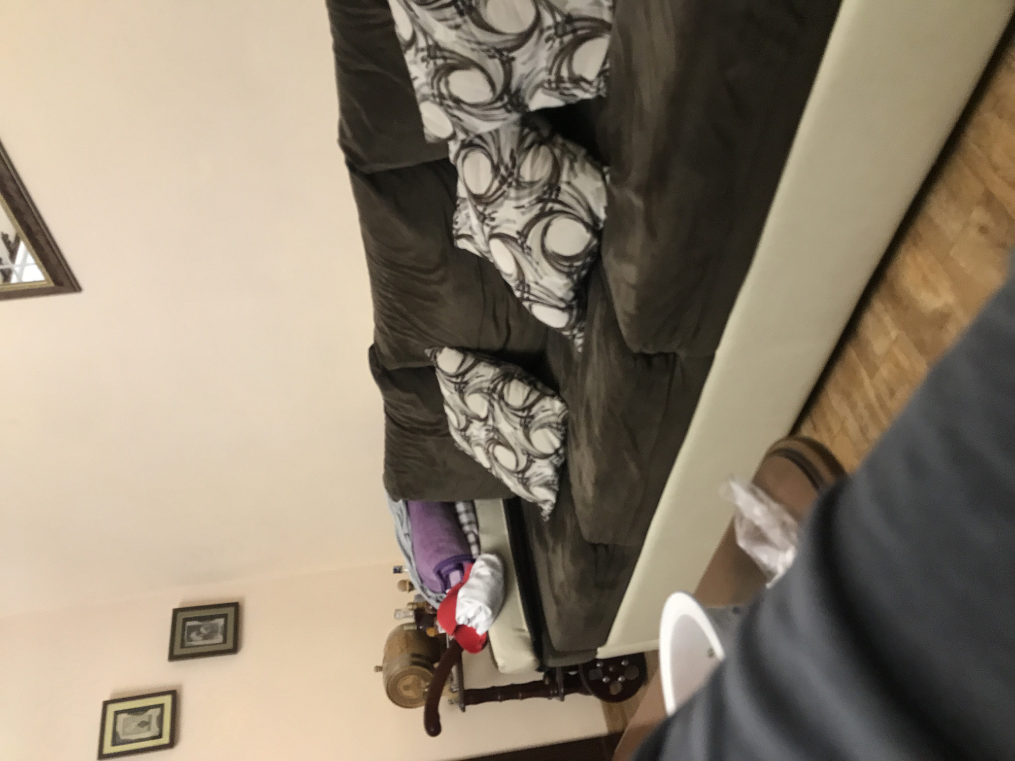 The image depicts a domestic interior. A brown sofa with patterned cushions is partially visible, along with a small wheeled table carrying items that appear to include a towel and small bottles. Two framed pictures hang on a peach-colored wall. The location is possibly Yerevan, Armenia. A person wearing a grey garment is partly in the foreground.