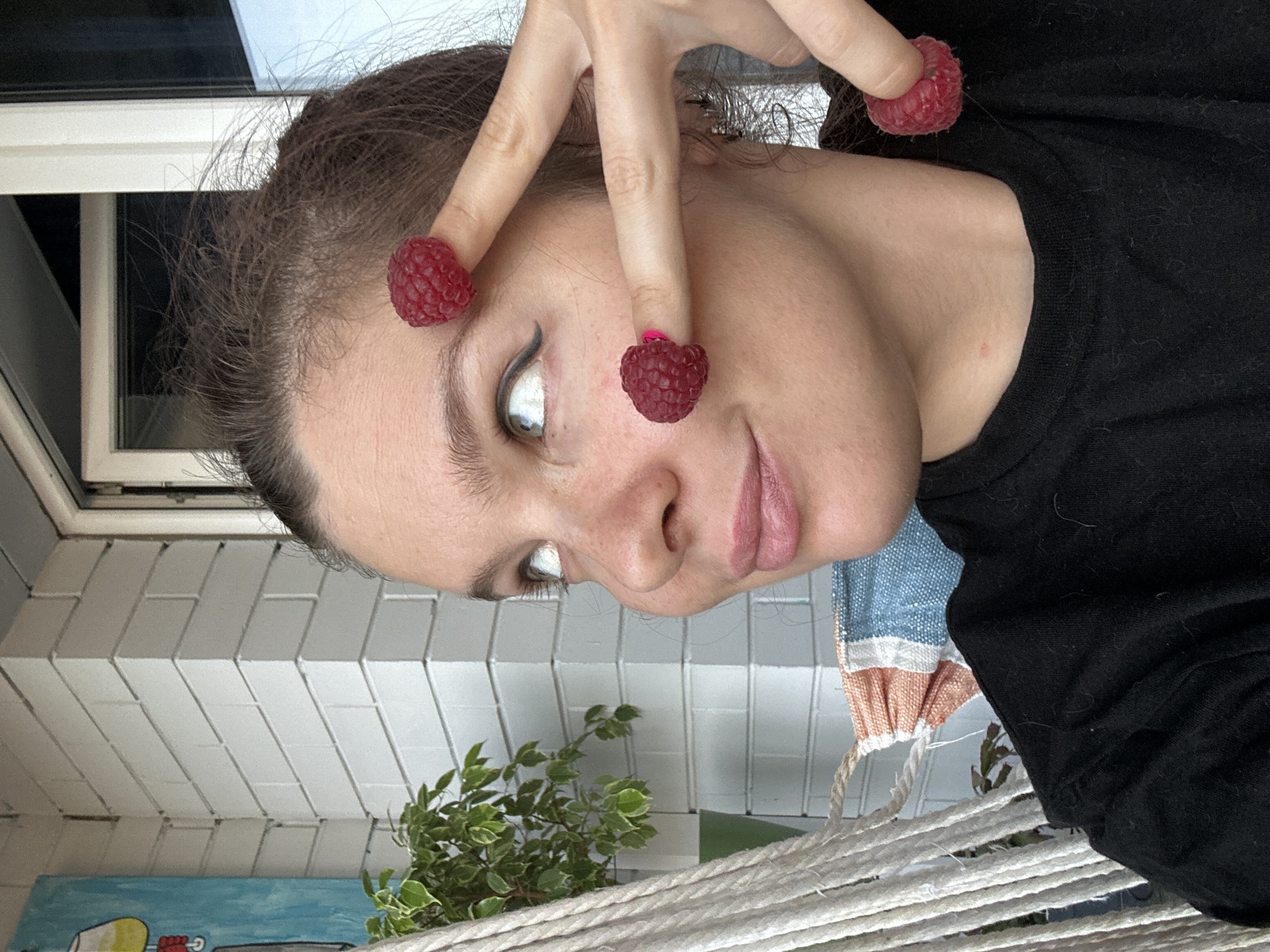 A young woman is depicted close-up, posing with fresh raspberries held to her fingers. She is wearing a black shirt and has visible makeup, including eyeliner. The location appears to be indoors, with white brickwork visible in the background, along with a plant and a painting. A window and parts of a hammock can be seen. There is no visible indication of the location being Vladimir, Russia.