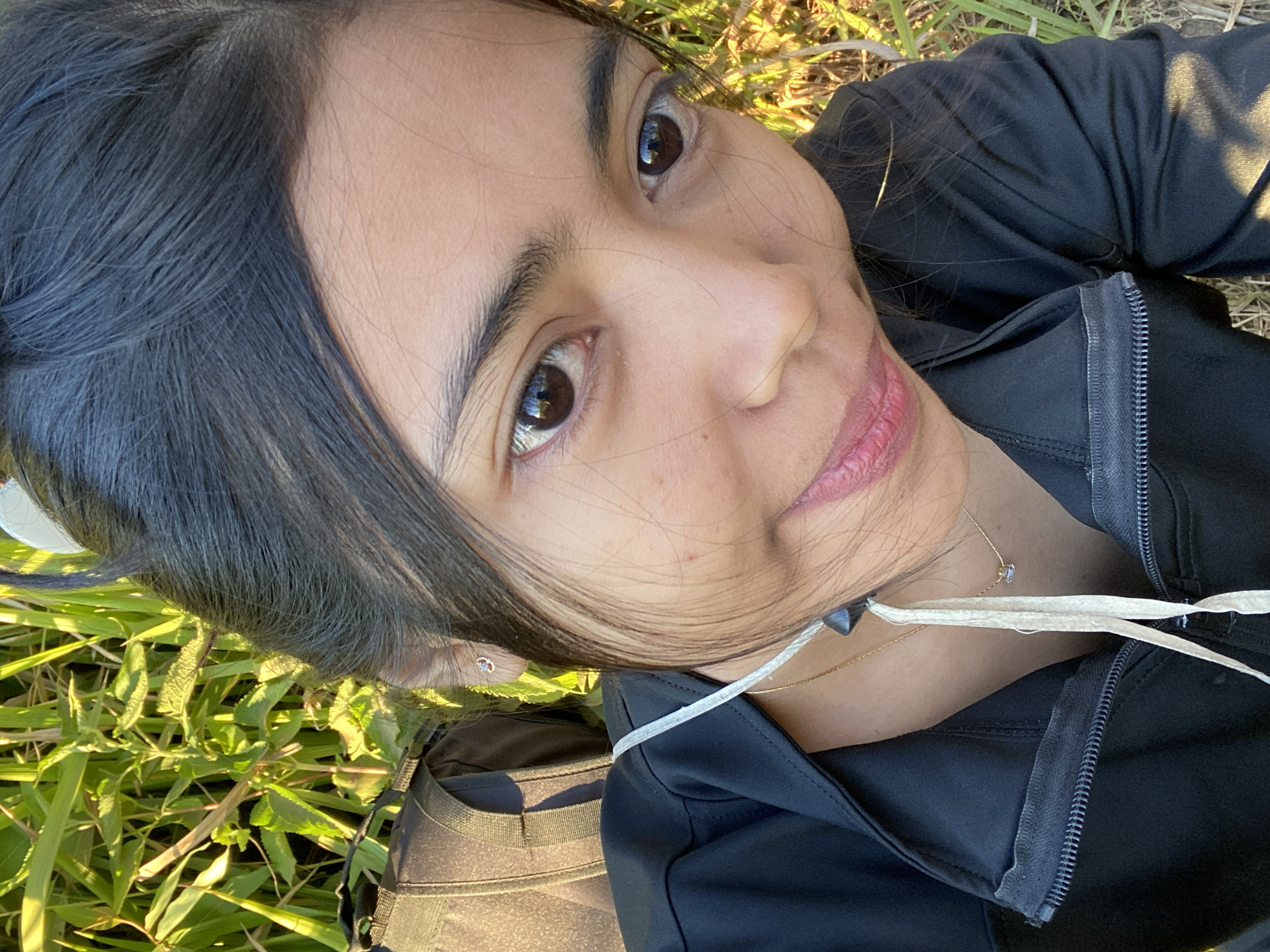 The image is a close-up, outdoor selfie taken in a grassy area with sunlight illuminating the scene. A young woman with dark hair pulled back is the primary subject. She is wearing a black jacket with a zipper and a thin gold necklace with a small pendant. A light-colored string hangs from her jacket, attached to dark objects near her neck. A dark backpack is visible behind her left shoulder. The background consists of tall green grass and vegetation, suggesting a natural environment. There is no discernible information about the city of Gondosuli, Indonesia, in the image itself.