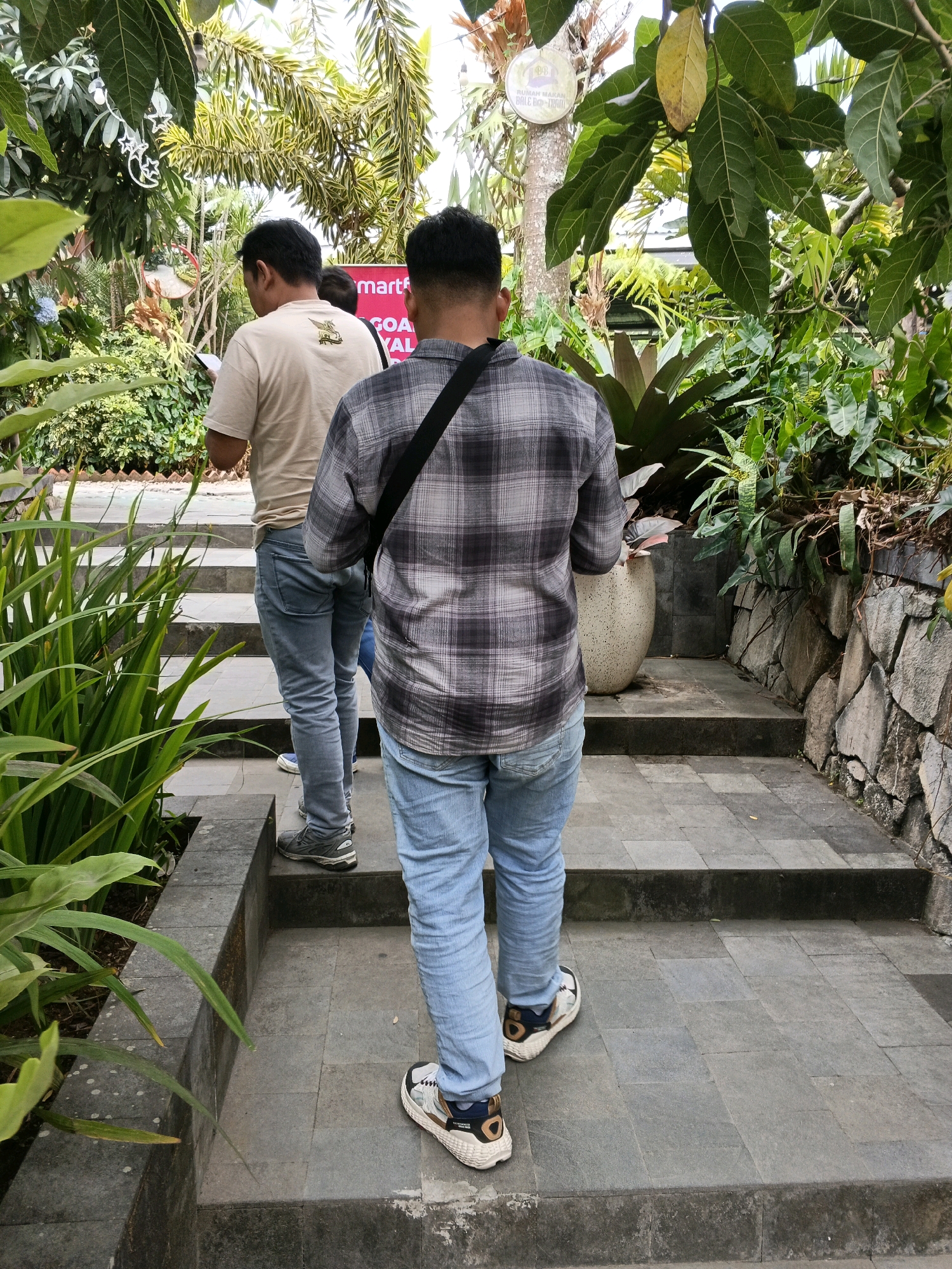 The image depicts two men walking up a stone staircase in an outdoor garden setting in Ngargoyoso, Indonesia. The man in the foreground, facing away from the camera, is wearing a grey and black plaid shirt and light blue jeans. He has a black strap across his chest. The second man, to his left and slightly ahead, is wearing a light beige t-shirt and light blue jeans. Both men appear to be holding or looking at something in their hands. The staircase is flanked by lush greenery, including large-leafed plants and trees. A stone wall is visible on the right side of the image, and a large decorative pot is placed near the staircase. In the background, some foliage and a sign are visible, suggesting a commercial or recreational area.