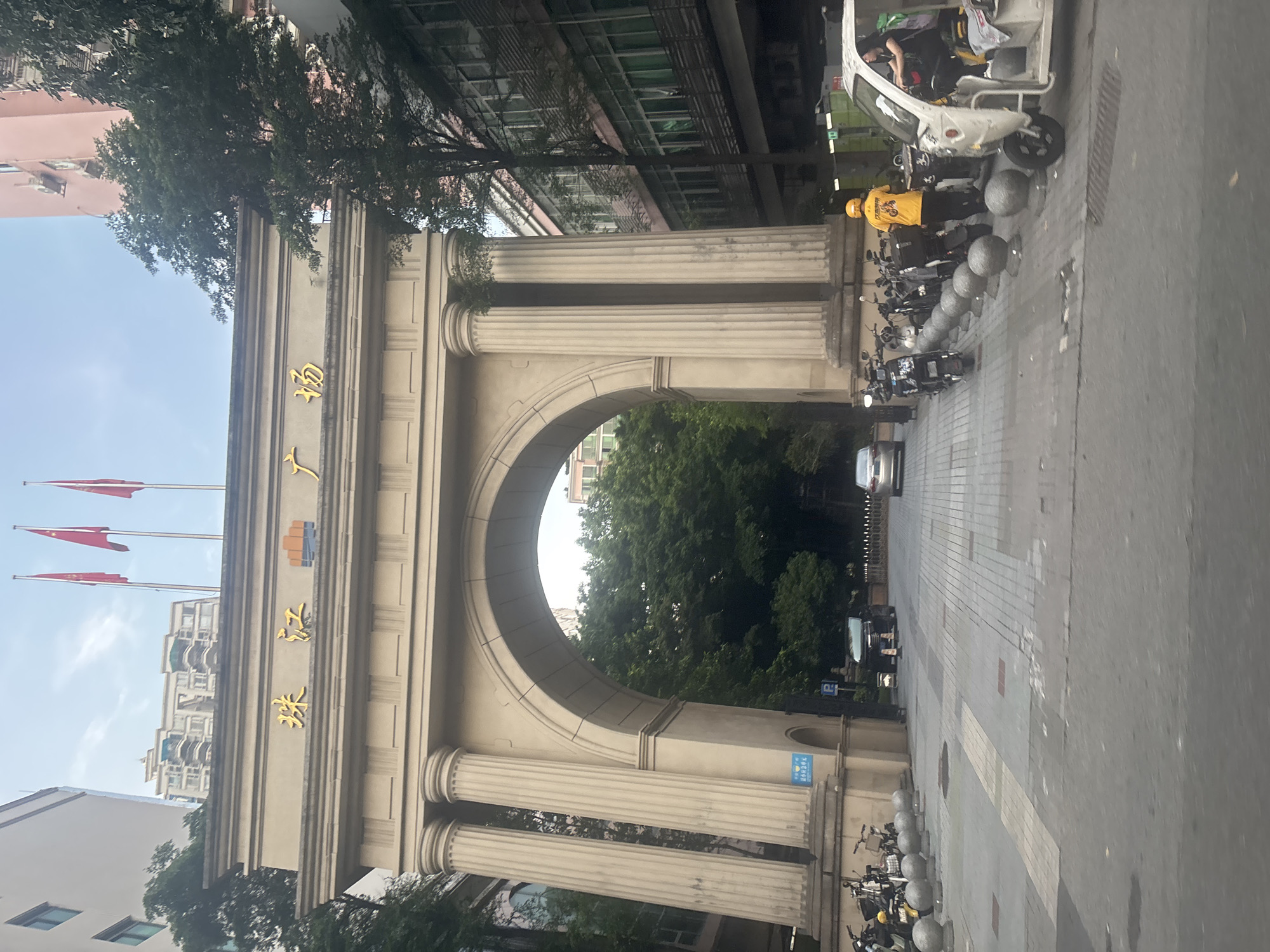 The image shows an ornate archway with Chinese characters inscribed at the top. The characters are in gold and appear to read "珠江广场" which translates to "Zhujiang Square". Three red flags are flying on flagpoles above the archway. The archway is supported by large, fluted columns and has a wide, arched opening.
Beyond the archway, trees and buildings are visible. In front of the archway, there are several parked motorcycles and scooters. A few people are present, including a person wearing a yellow uniform and helmet, possibly a delivery driver, standing near a white three-wheeled vehicle. Another person is seated in the white vehicle. The ground is paved with interlocking bricks. The scene appears to be a public square or entrance in Haizhu District, China. The overall lighting suggests it is daytime, possibly late afternoon or early evening.
