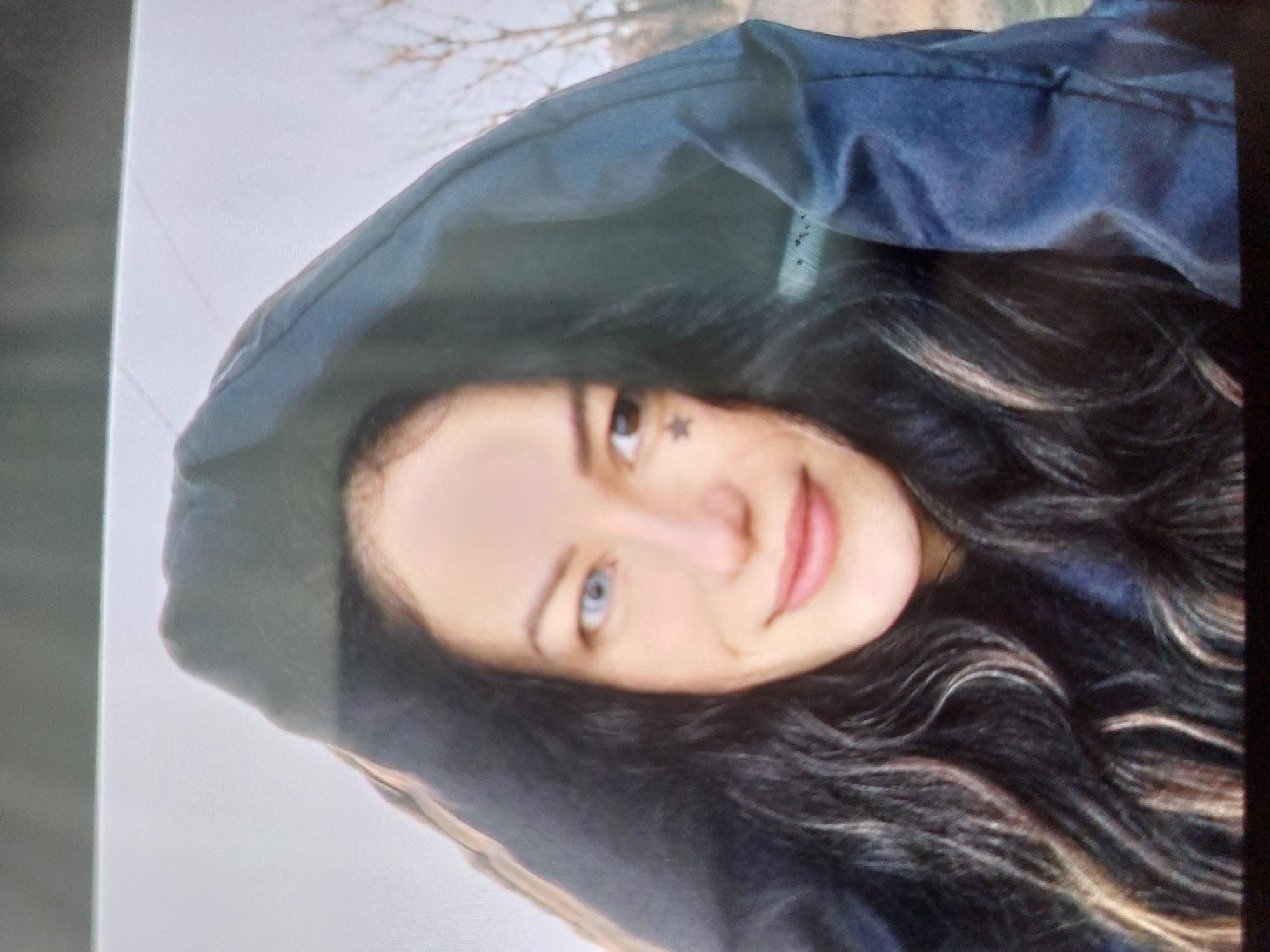 A woman with long, dark hair and fair skin is wearing a dark blue hooded jacket. The hood is pulled up, partially framing her face. She has heterochromia, with one blue eye. A small star tattoo is visible near her eye. The background appears to be a blurred outdoor scene with bare trees. The image might have been taken in Tsarevo, Bulgaria, but there are no visual cues to confirm the location.