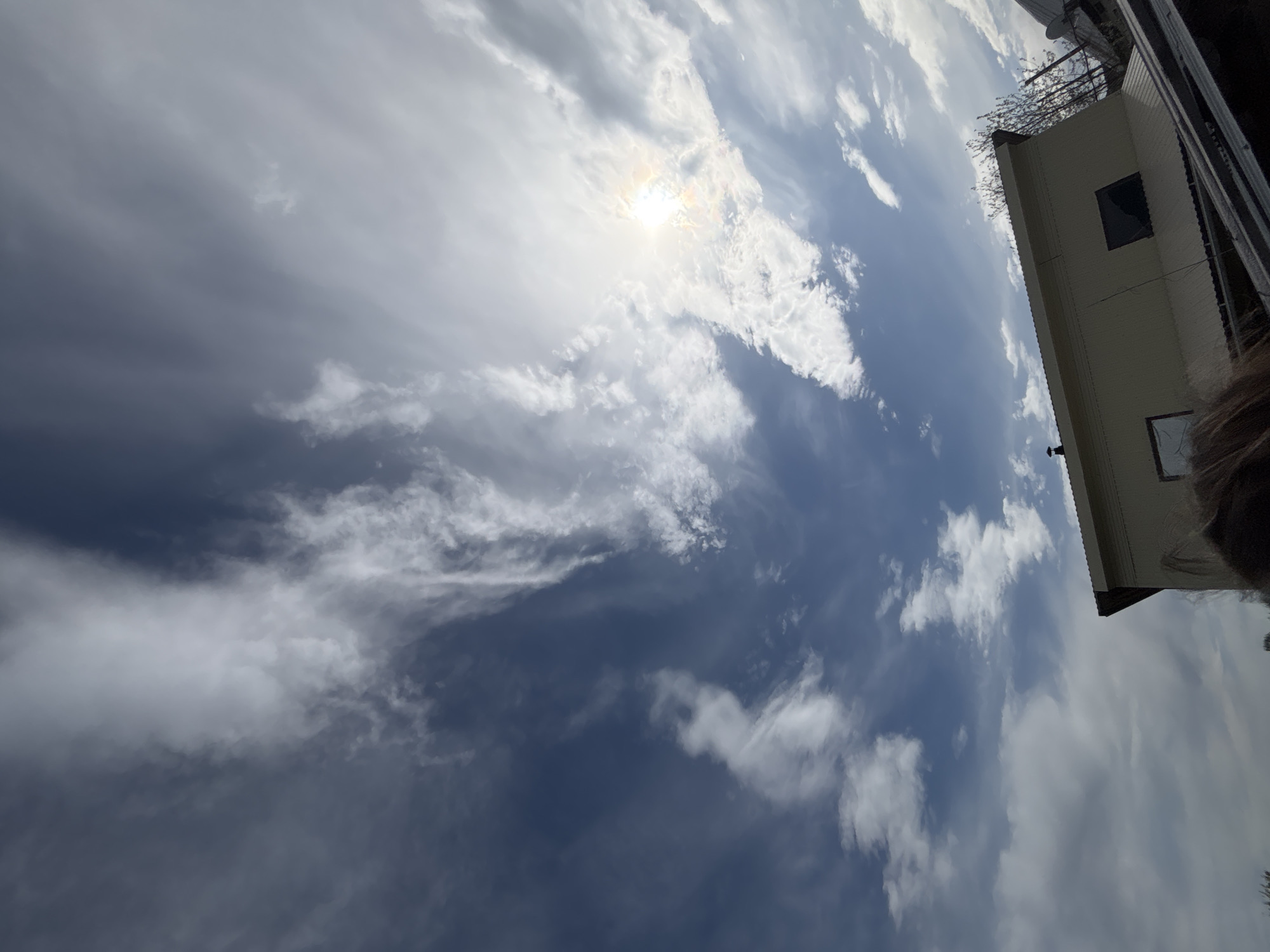 The image presents a daytime scene under a sky partially obscured by clouds, with the sun shining through. In the upper-right corner, a corner of a light-colored building is visible, possibly a residential or commercial structure. The sky transitions from a bright, sunlit area to darker, cloudier patches. The location is presumably in Фоки, Russia.