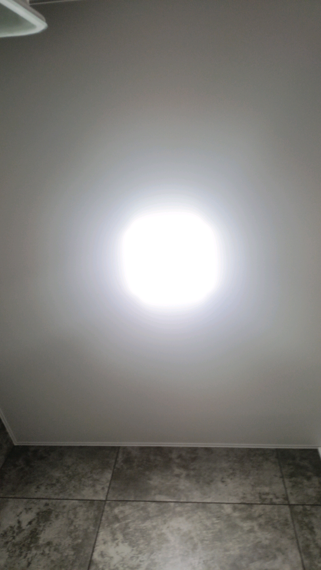 The image depicts a close-up, low-angle shot of a ceiling with a bright light fixture at its center, emitting a strong, diffused glow. The ceiling appears to be made of a smooth, white material. Below the ceiling is a tiled surface, likely a wall or floor, with large, square tiles in shades of gray and black, exhibiting a mottled, stone-like pattern. The overall lighting suggests an indoor environment, possibly a bathroom or a modern interior space. Without additional information or context, it is impossible to determine the specific location within Homyel, Belarus.