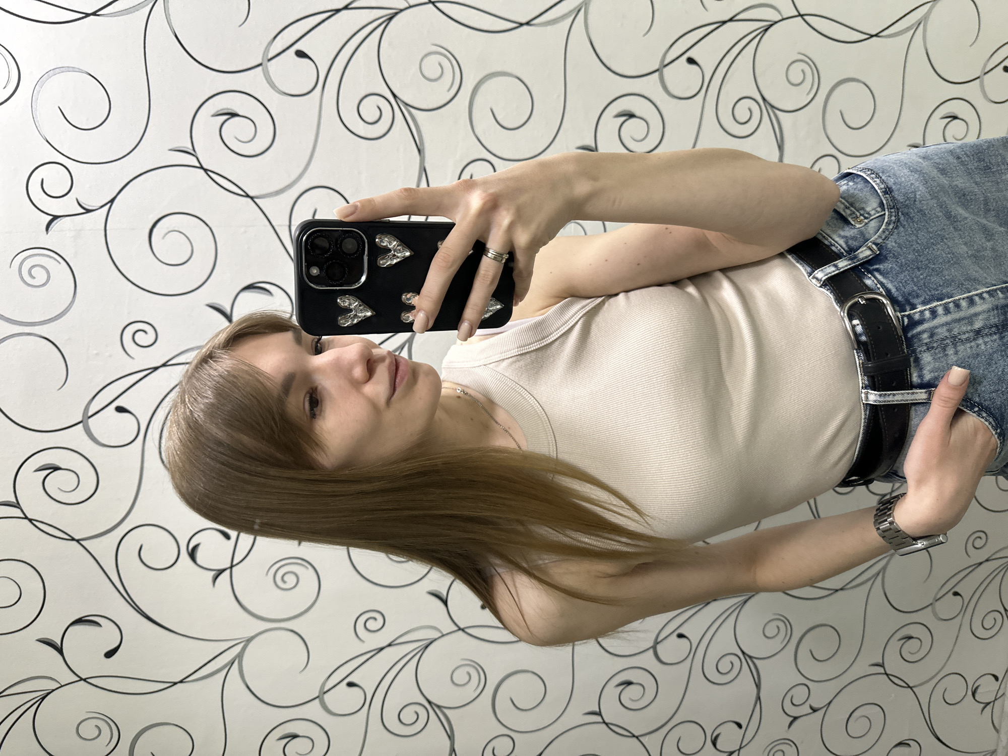 The image shows a woman taking a selfie indoors. She is holding a black smartphone with silver heart embellishments on the back, showcasing the camera lenses. The woman has brown hair with bangs and is wearing a light-colored tank top, jeans with a black belt, and a watch on her left wrist. A simple ring is visible on her finger. The background is a wall covered in a black and white swirling pattern. The location, Chaikovsky, Russia, is not discernible from the visual information.