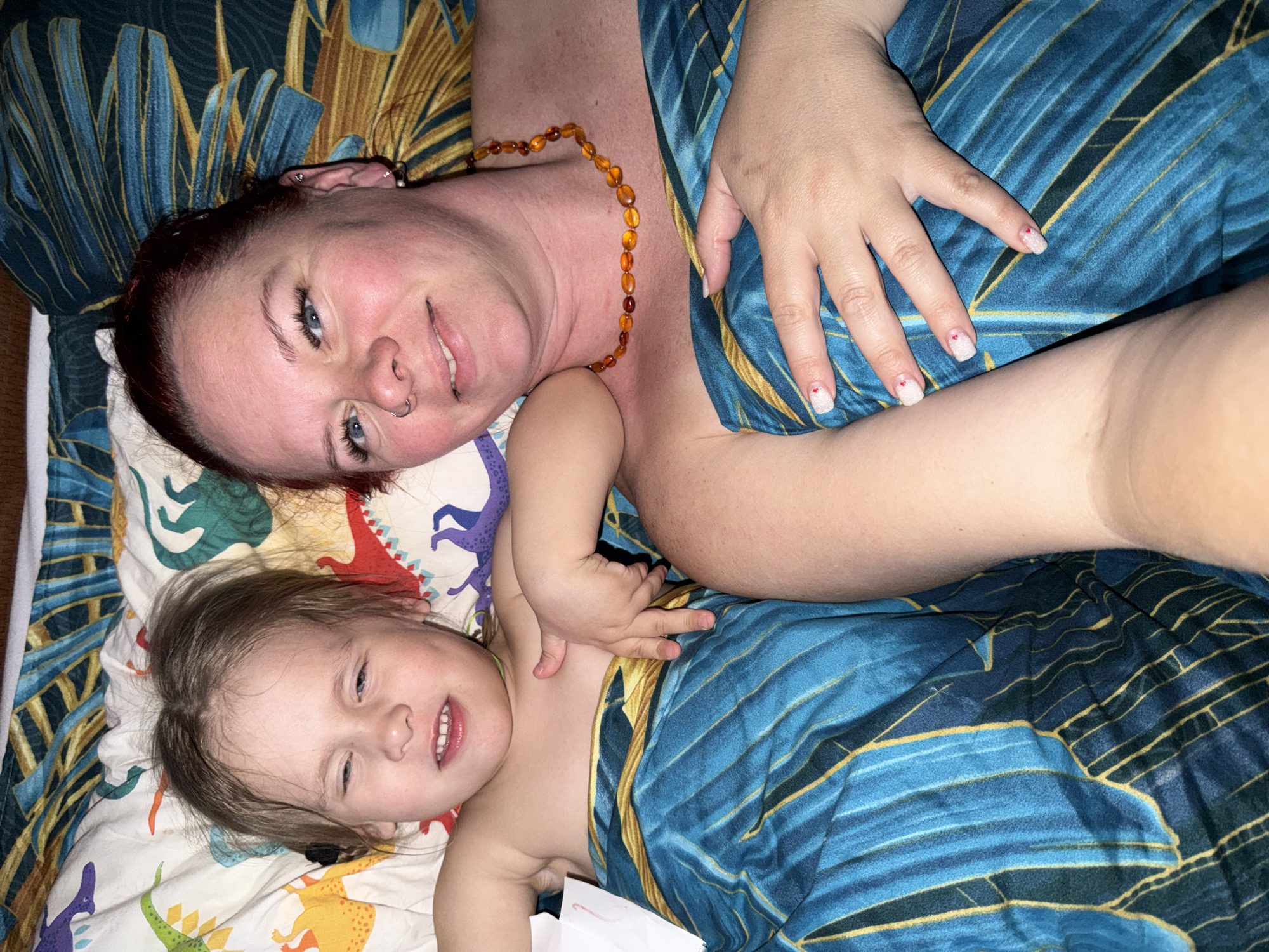 The image depicts an adult woman and a young child lying on a bed. They are both partially covered by a blue and gold patterned blanket. The woman is wearing an amber beaded necklace and has a nose ring. She has fair skin and reddish hair. Her nails are painted with a white and red heart design. The child has fair skin and blonde hair. The bed is covered with a dinosaur-themed patterned sheet. The location is unknown.