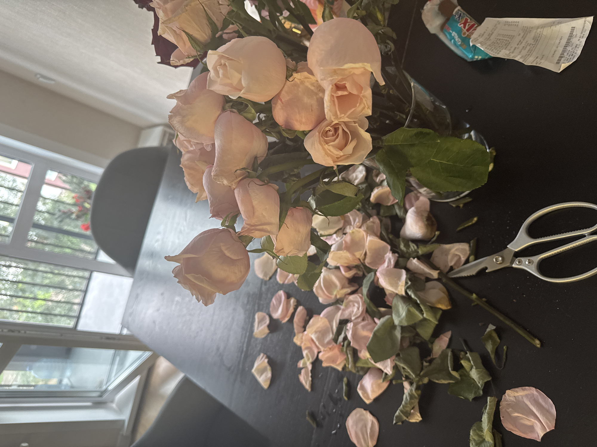 The image depicts a close-up, high-angle shot of a dark wooden table in what appears to be an indoor setting. On the table, a bouquet of wilted pink roses is arranged in a clear glass vase. Scattered around the vase are numerous fallen rose petals and leaves, suggesting the flowers are past their prime and possibly being processed or discarded. A pair of small silver scissors lies on the table near a discarded rose stem. To the right of the vase, a crumpled receipt is visible, partially obscured by a blue and white wrapper. In the background, a window with a view of greenery and buildings is partially visible, along with furniture that suggests a domestic or office environment. The lighting suggests it is daytime. The scene appears to be located in the city of Perm, Russia, given the context of the prompt, although this cannot be definitively confirmed from visual elements alone.