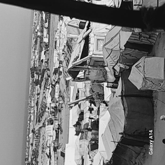 The black and white image portrays a dense slum or refugee camp. Makeshift dwellings are constructed from various materials like cloth, wood, and metal, creating a chaotic, overcrowded environment. Structures are closely packed, indicating high population density. The horizon shows similar constructions stretching far into the distance. The photograph's overall tone suggests poverty, displacement, and hardship.