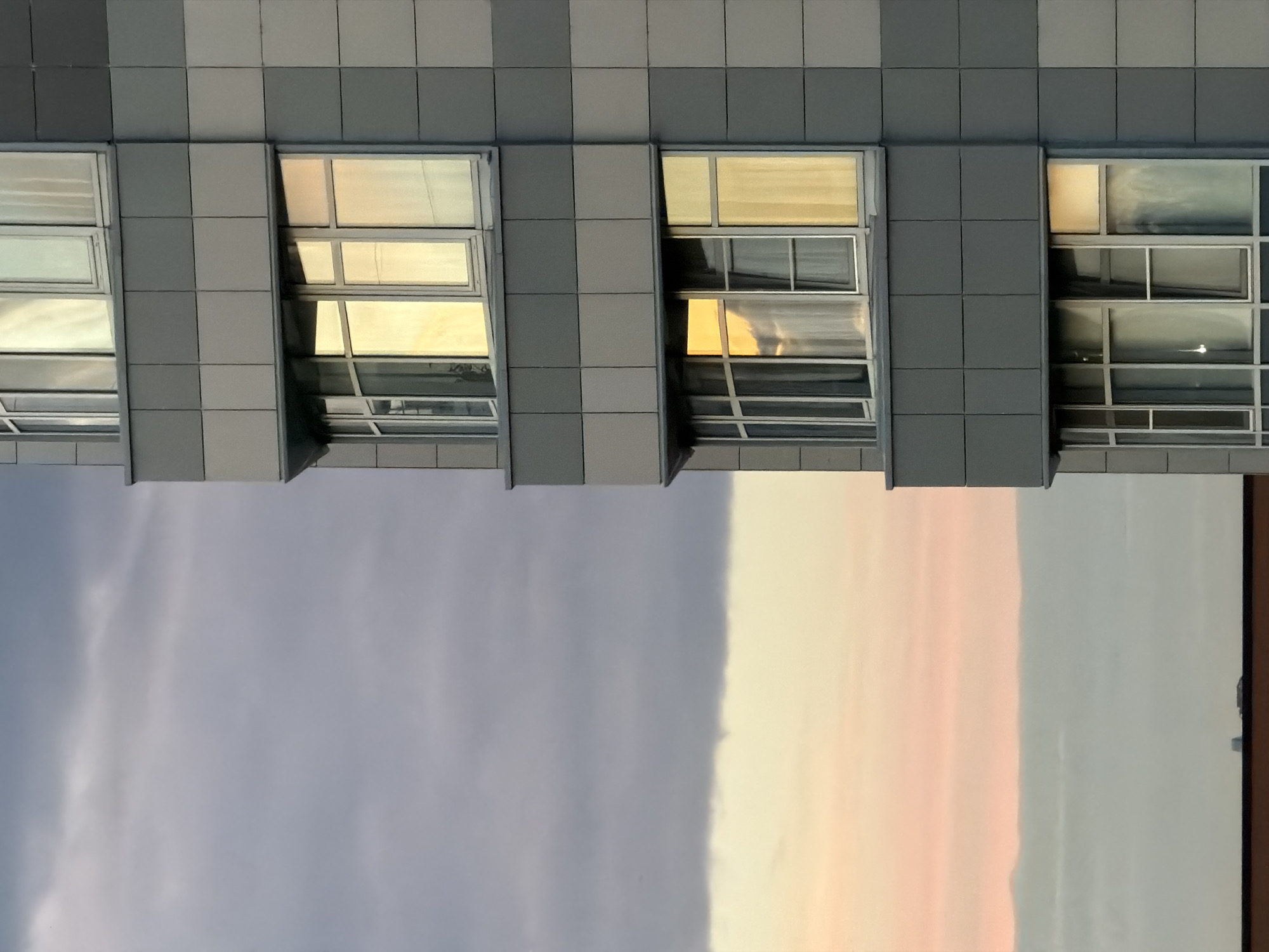 The image captures a close-up, angled view of the exterior of a modern building, likely an apartment complex, on the right side, juxtaposed with a muted sky on the left. The building's facade is composed of large, square panels in varying shades of gray, giving it a geometric, tiled appearance. Several windows are visible, some reflecting the warm hues of what appears to be a sunset or sunrise. The windows are framed in silver and contain white curtains. The sky transitions from a light pink and yellow at the bottom to a darker gray with visible cloud formations towards the top. A sliver of a darker, solid structure, possibly a rooftop or another building, is visible at the very bottom edge of the frame. The scene appears to be devoid of human presence.