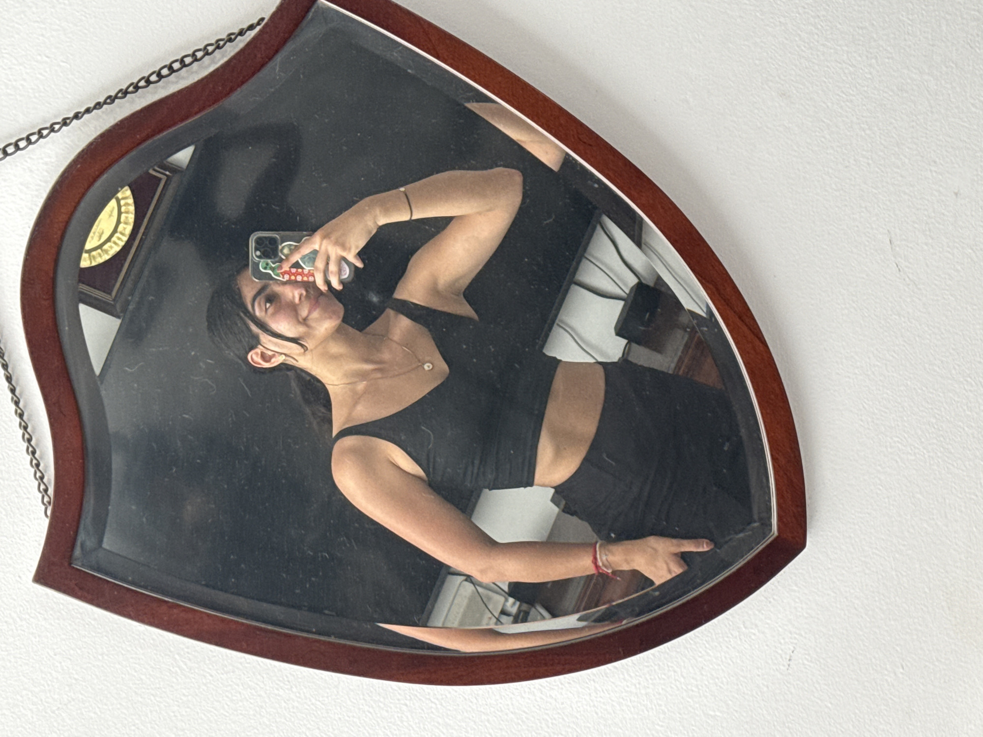 A young woman is depicted in a mirror selfie, captured within a heart- or shield-shaped mirror featuring a reddish-brown wooden frame and a dark metal chain for hanging. She smiles broadly at her reflection, holding a smartphone with a colorful, patterned case. She is wearing a black crop top that reveals her midriff and black bottoms, likely athletic wear. Her dark hair is pulled back, with some strands framing her face, and she has a small stud earring. Accessories include a delicate necklace with a round, light-colored pendant, a black bracelet on one wrist, and a red and white braided bracelet on the other.
The mirror's reflection offers glimpses of the room behind her, including a vintage-style wall clock with a golden face and dark frame, and what appears to be dark furniture or possibly a bed with striped bedding. The wall outside the mirror is white and textured, with minor marks. The overall mood is casual and cheerful. No discernible text is visible on the phone case or other elements. The location context is Puebla, Mexico.