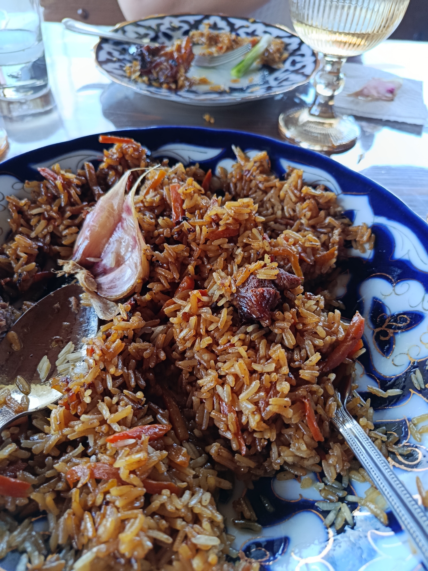 A close-up, eye-level shot focuses on a large blue and white decorative plate filled with pilaf. The pilaf is a rich, golden-brown color, composed of rice, shredded carrots, and chunks of meat. A whole head of garlic, partially cooked and peeled, is prominently placed in the center of the dish, resting on top of the pilaf. Two spoons, one silver and one with a patterned handle, are partially submerged in the pilaf, suggesting recent consumption. The plate is set on a wooden table, with a clear glass of white wine in a stemmed glass visible in the background to the right. To the left, another glass of clear liquid and a plate with leftover food and a fork can be seen. The scene appears to be indoors, possibly in a restaurant or dining room, with soft lighting. There are no visible individuals in the frame. The overall impression is of a meal being enjoyed, with the focus sharply on the pilaf.