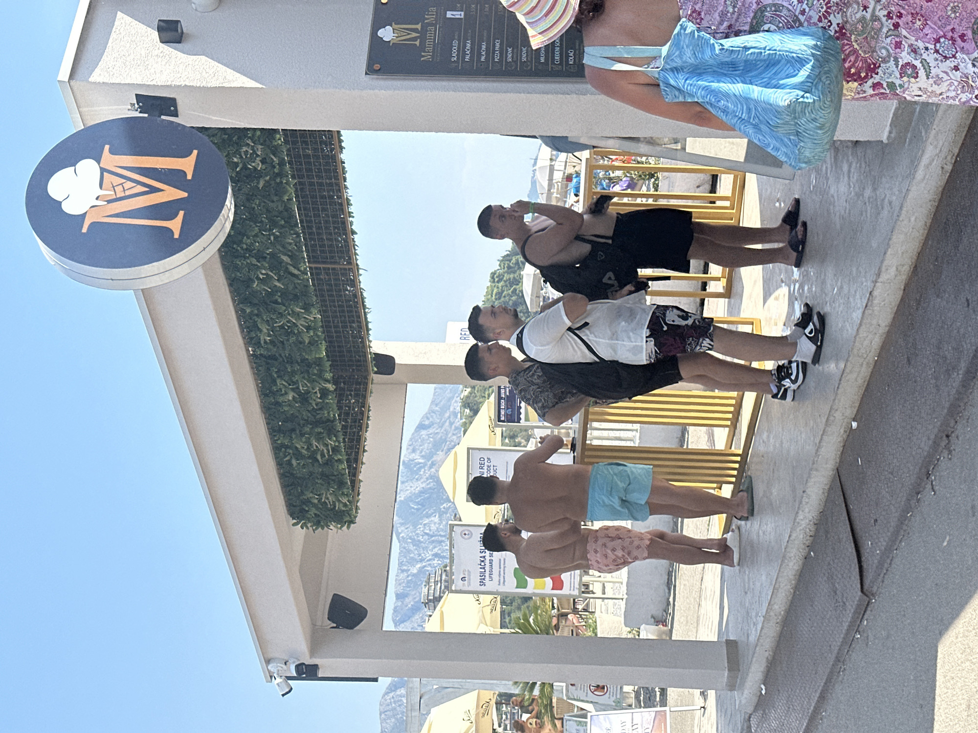 In Budva, Montenegro, a group of men, appearing to be in their twenties, stand in a queue outside an establishment with a prominent "Mamma Mia" sign featuring an ice cream cone logo. The men are dressed casually in beach attire. The establishment is located on a street adjacent to what appears to be a beach or promenade, with the Adriatic Sea and mountainous terrain visible in the background. The scene is bathed in bright sunlight under a clear blue sky. One man in the foreground is wearing a colorful striped sun hat and a floral-patterned dress. There are several white beach umbrellas visible further down the street, suggesting a tourist area.