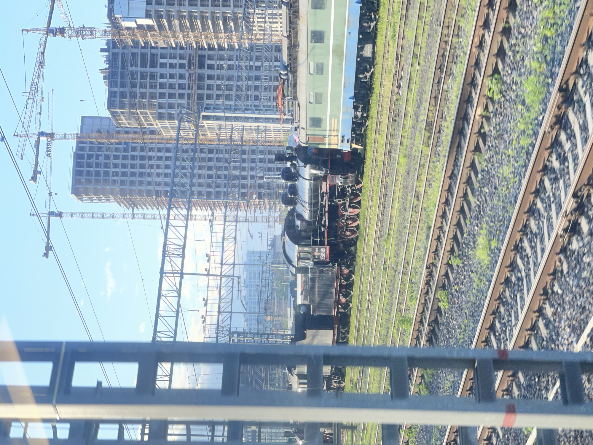 The image shows a steam train on a railway track, with modern high-rise buildings and construction cranes in the background. The photo appears to have been taken from a bridge or elevated structure, as indicated by the metal railing in the foreground. The location is Khimki, Russia, identifiable by the architectural style of the buildings under construction. The steam train is black, with visible red wheels and a green and blue carriage attached. The train is moving along multiple parallel railway tracks. The overall environment suggests a juxtaposition of old and new, with traditional railway transport alongside modern urban development. The sky is pale blue with minimal clouds.