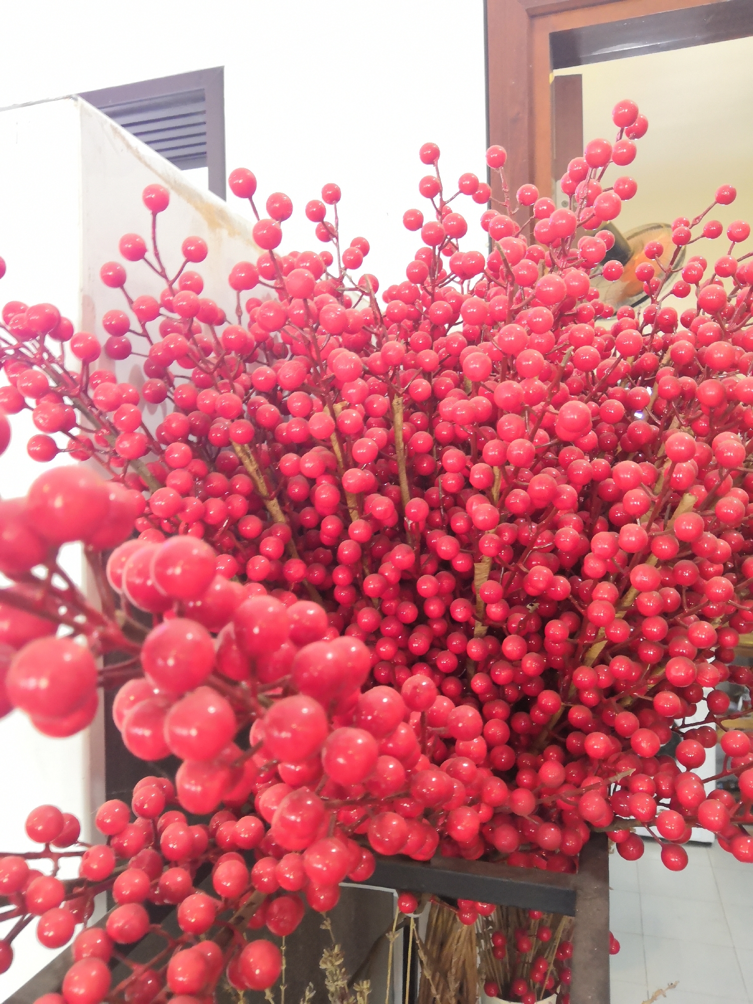 A close-up shot focuses on a dense arrangement of artificial red berries on thin, brown stems. The berries are round and shiny, clustered together to form a vibrant, textured mass. In the background, a white wall with a dark-slatted element is visible on the left. To the right, a wooden-framed doorway is partially seen, with a reflection of a ceiling fan and what appears to be another cluster of red berries in the mirror. A dark metal shelf or stand is positioned in front of some of the berry branches, with additional dried branches and berry stems visible below. The scene suggests an indoor decorative arrangement, possibly in a shop or home in Bac Nha Trang Ward, Vietnam.