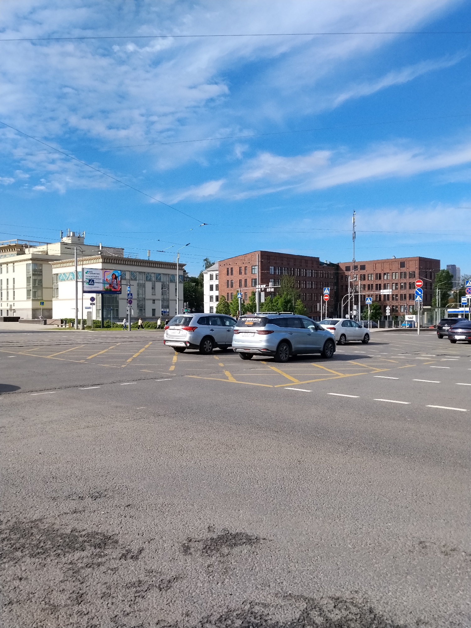 The image depicts a cityscape in Moscow, Russia, during daytime under a partly cloudy sky. A major road intersection dominates the foreground, marked with yellow grid lines and white lane dividers on an asphalt surface showing wear. Several cars are present: a silver Mitsubishi Outlander and a silver SUV, possibly a Haval F7x, are positioned centrally. Further vehicles are discernible in the background. Two buildings are visible: on the left, a white building with glass elements and signage; on the right, a larger red brick building. Pedestrians can be seen crossing at designated areas near the white building. Traffic signs are mounted on poles near crosswalks. Overhead power lines traverse the sky.