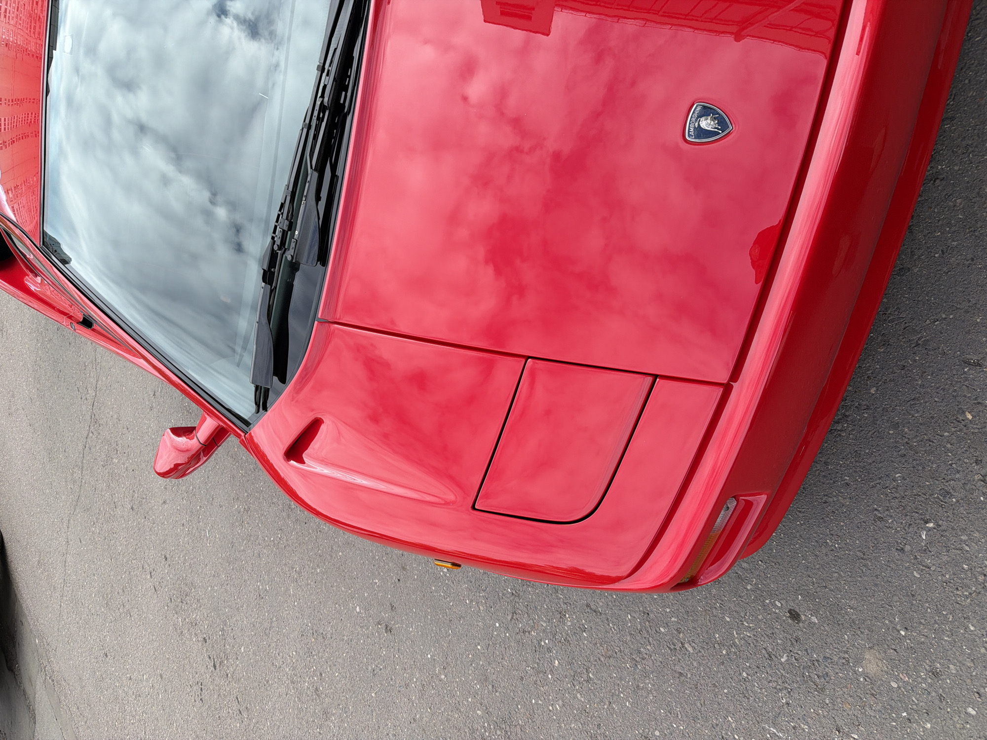 An overhead, slightly angled shot captures the front section of a vibrant red sports car, likely a classic model given its design, parked on an asphalt surface. The vehicle's highly polished, glossy paint reflects the cloudy sky above, creating a distorted yet clear image of cumulus clouds across its hood and windshield.
The primary subject is the car itself, which dominates the frame. Its striking red color is consistent across its body panels. On the front hood, a silver-edged emblem featuring a charging bull is prominently displayed, with the word "LAMBORGHINI" visible around its perimeter. Below this, the car's design features a rectangular pop-up headlight cover, suggesting an older, iconic sports car model. A small, amber-colored turn signal is visible near the front bumper on the right side.
The windshield reflects a mostly overcast sky, with distinct cloud formations and patches of brighter sky. Windshield wipers are positioned at the base of the glass. To the far left, a red side mirror and a portion of the car's door are visible, also reflecting elements of the surroundings. The ground beneath the car is dark gray asphalt, textured with small aggregate stones and some faint cracks. There is no human activity or event taking place; the scene depicts a stationary vehicle outdoors during daylight hours under a cloudy sky. The location context is Moscow, Russia.
