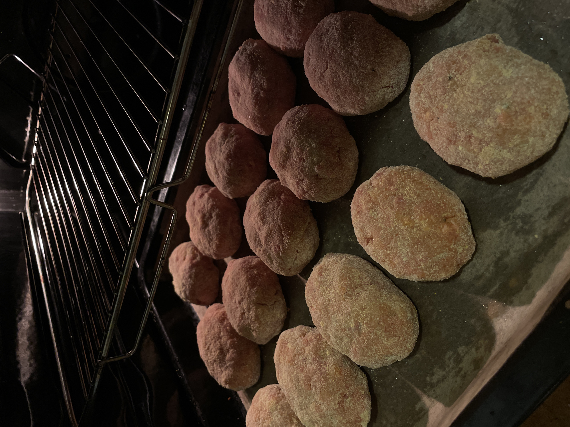 The image shows a batch of raw, breaded cutlets or patties arranged on a baking sheet, partially inside an oven. The cutlets are oval-shaped and covered in a light-colored breading, with visible specks of seasoning. They are densely packed on the baking sheet, suggesting they are being prepared for cooking. To the left of the baking sheet, the metallic racks of an oven are visible, illuminated by the oven light, indicating the setting is likely a kitchen. The overall lighting is dim and warm, suggesting this might be an evening scene or the interior of the oven. There is no visible text in the image.