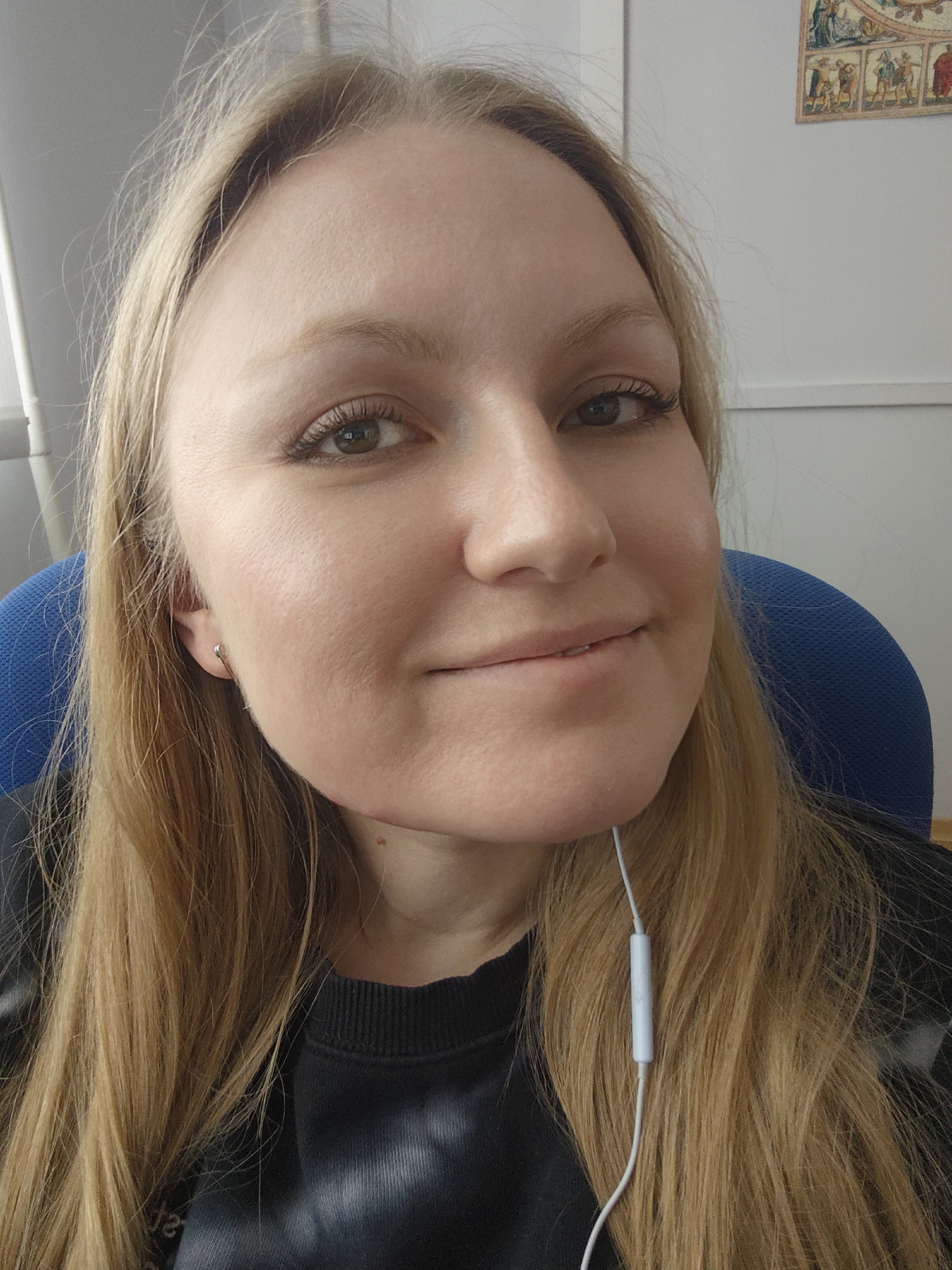 A fair-skinned woman with light brown hair is centered in the image. She is wearing a black sweater and has white earphones hanging around her neck. She is sitting in a blue chair. Behind her is a white wall, with a framed picture hanging on it. There is no information to indicate that the location is in the city of Novokuybyshevsk, Russia.