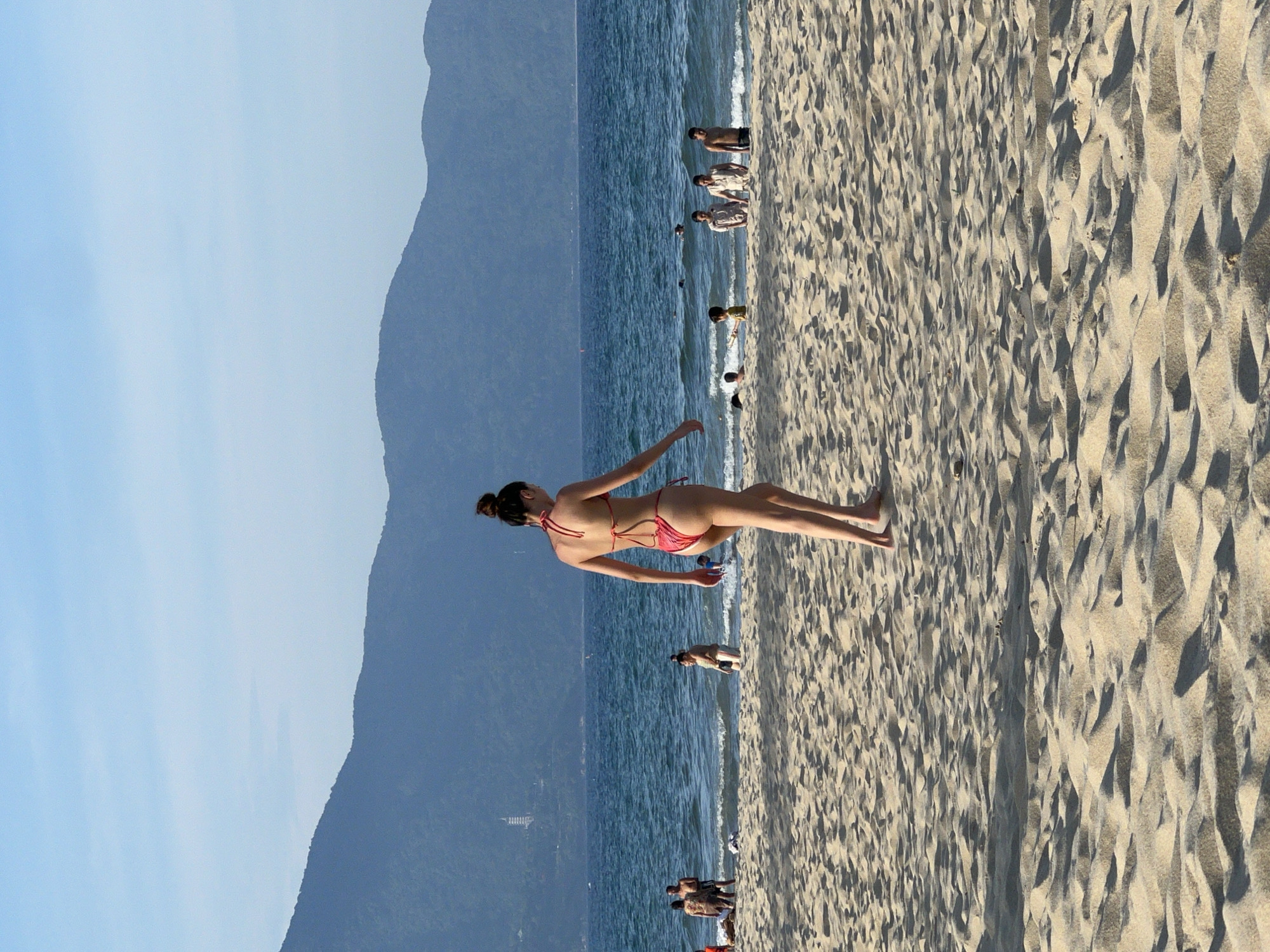 This is a photograph of a beach in Da Nang, Vietnam, with a woman in a red bikini walking away from the camera on the sand. She has her hair pulled up in a bun and is facing the ocean. In the background, the sea is visible with waves, and several other people are in the water, some swimming and others standing. Further in the distance, there are mountains, and a pagoda is visible on the slope of one of them. The sand is light-colored and textured with footprints. The sky is clear and blue.
