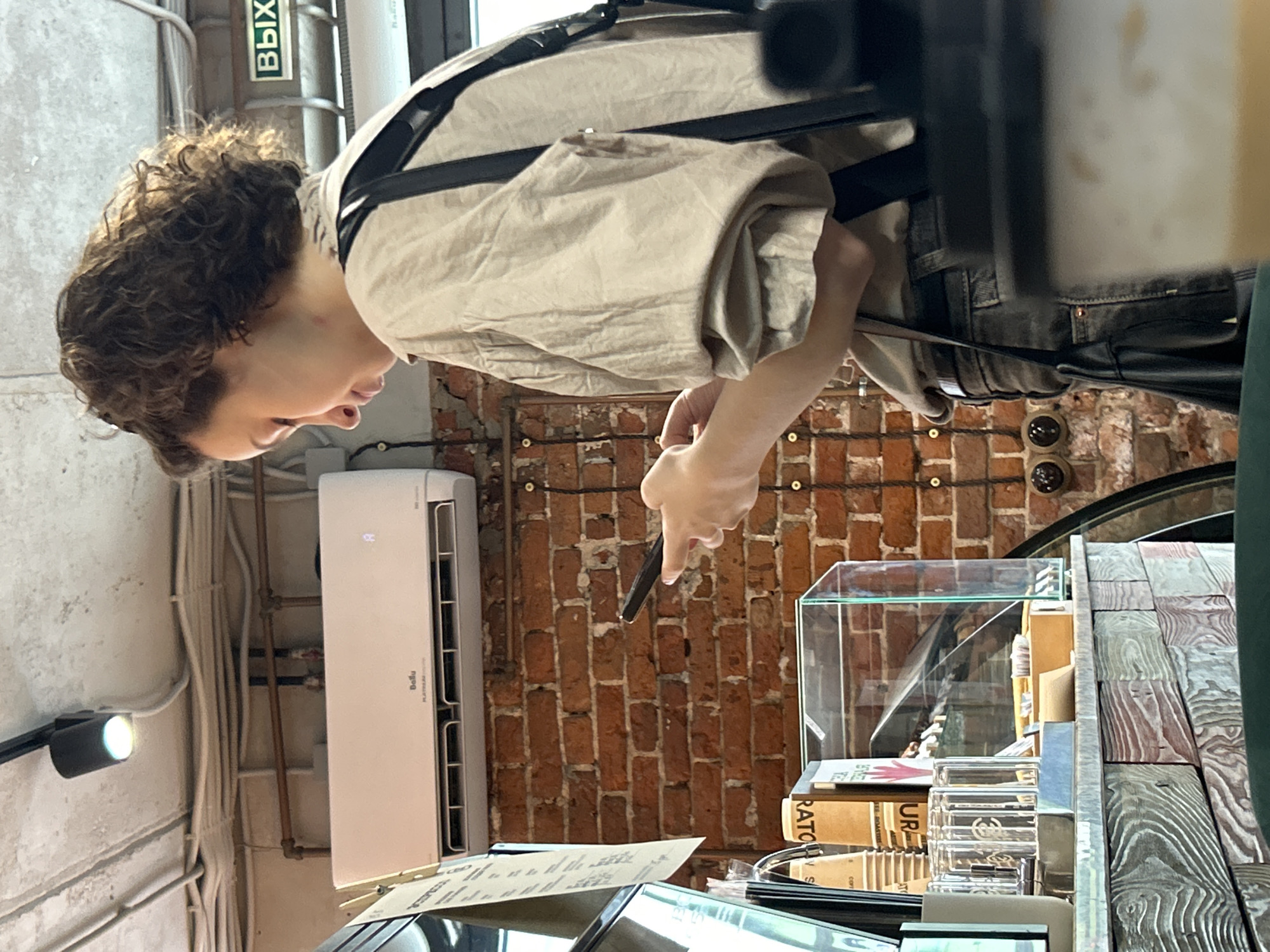 A medium shot shows a person holding a black pen over a counter. The person has short, curly brown hair and wears a tan shirt with black straps. The counter is covered in a wooden surface and glass display cases. Behind the person is a red brick wall. To the left of the person is a white air conditioning unit attached to the wall. Wires and pipes run along the wall near the AC unit. A black light fixture is also visible on the left side of the image.