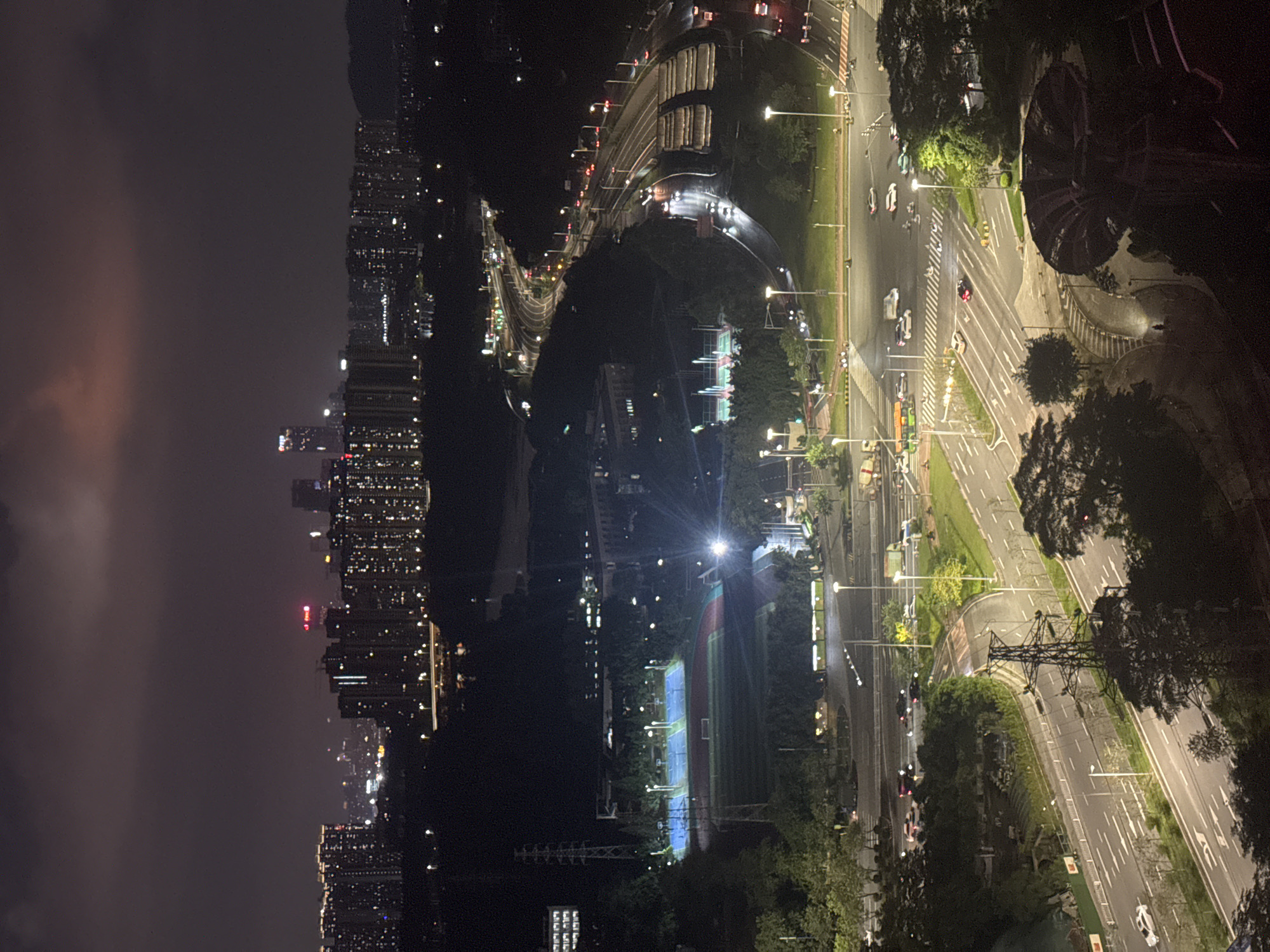 The image depicts a nighttime scene in the Panyu District of China, characterized by city lights and infrastructure. The left side shows a dense urban area with numerous buildings illuminated by countless lights. A highway winds through the area, with vehicles moving along it, their headlights visible. A sports complex with a running track and a football field is brightly lit. The right side of the image shows a network of roads with streetlights and moving cars. Trees line the roadways. An electrical tower is visible near the roadside. The sky appears dark with some faint, pinkish clouds near the cityscape.