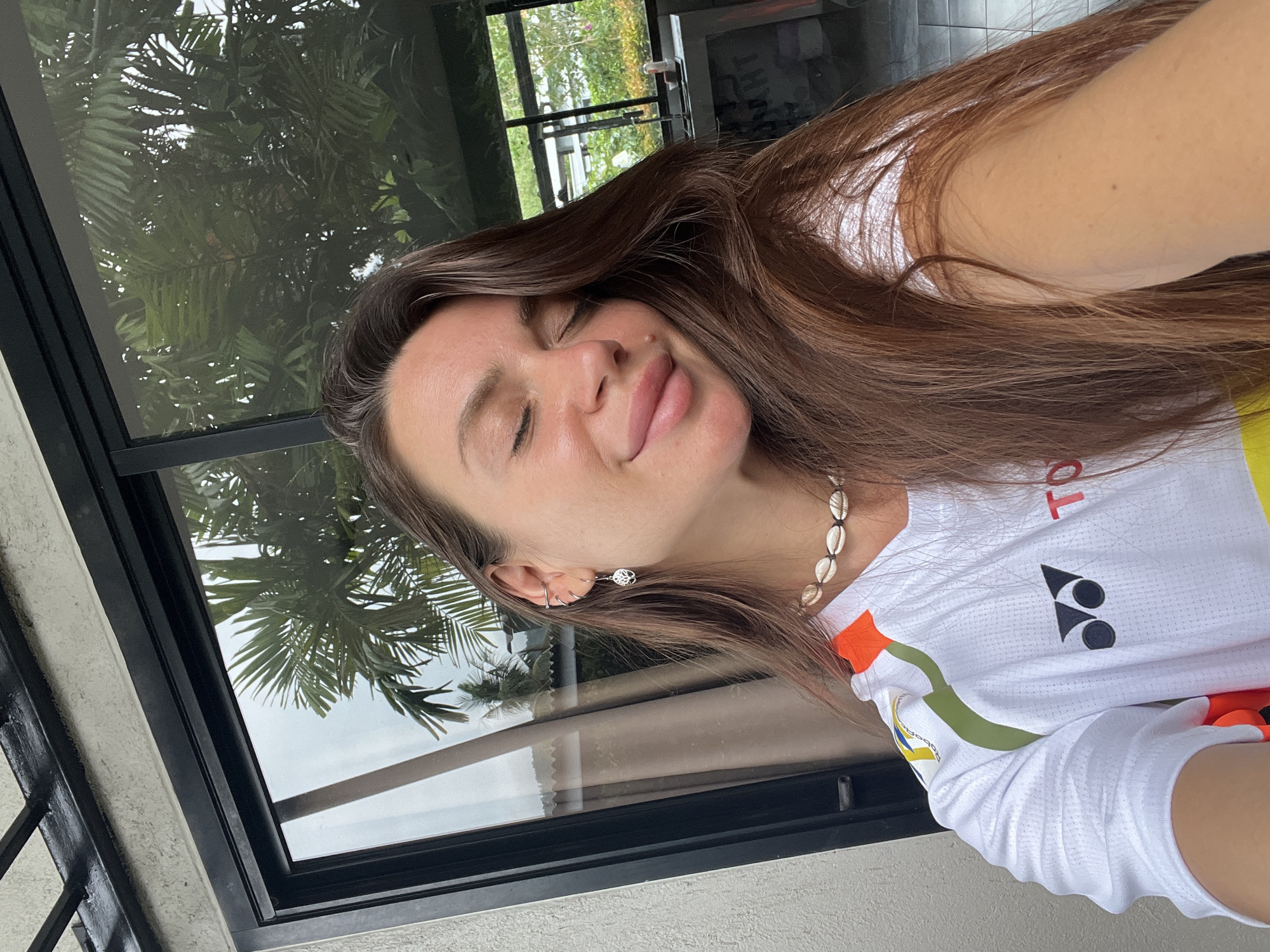 A woman with long brown hair is seen from the chest up, in an outdoor setting, with a window behind her. She has her eyes closed and is sticking out her tongue slightly. She is wearing a white athletic shirt with black and red logos and orange and green accents, along with a seashell necklace and earrings. Outside the window is a palm tree and foliage. The location is the city of Unknown, Unknown.