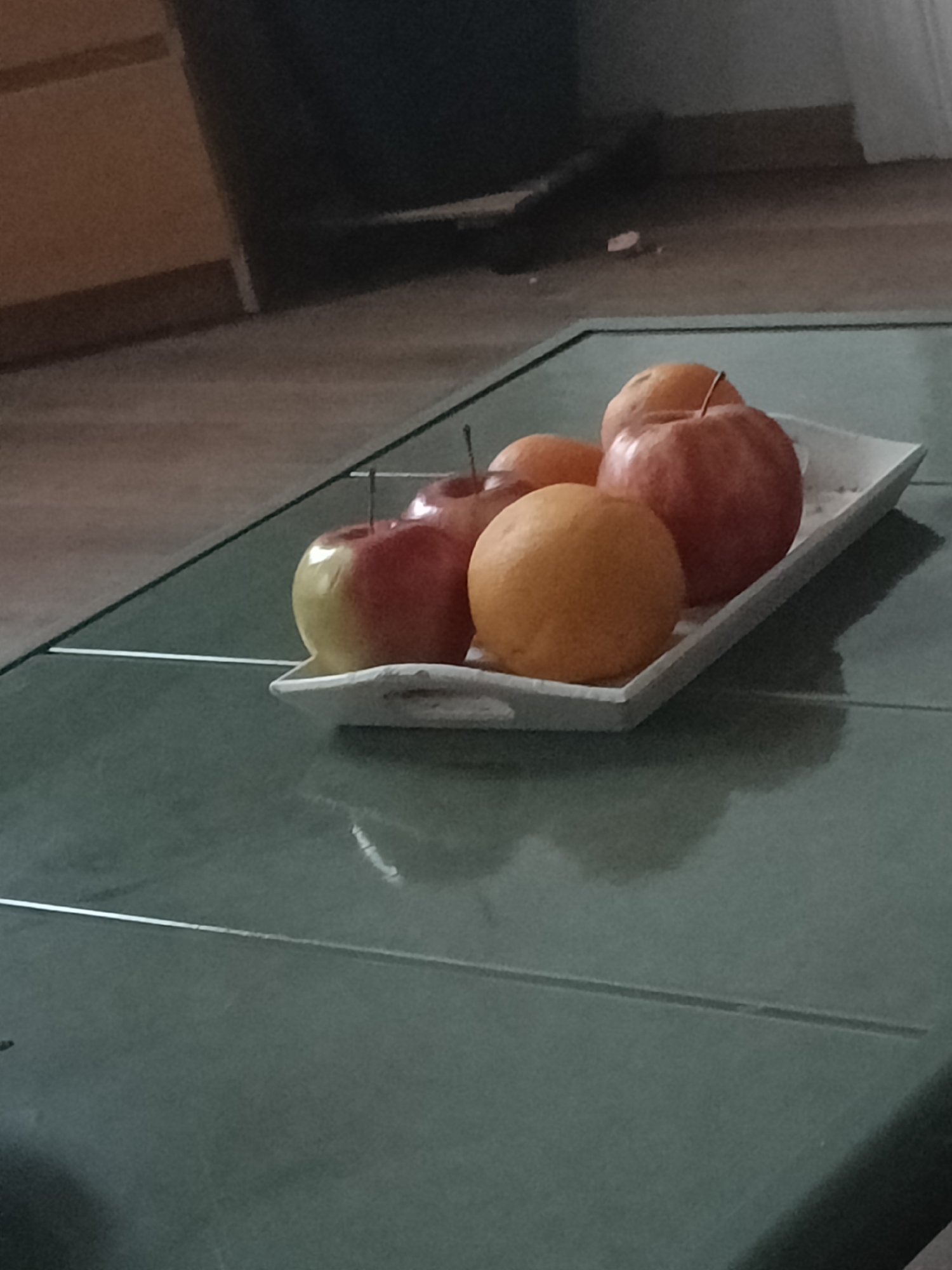 An indoor, eye-level shot captures a still life composition of fruits arranged on a white rectangular tray. The tray, positioned on a green-tinted glass table, holds five pieces of fruit: three apples and two oranges.
The apples are notable for their bicolored appearance, transitioning from red to yellow, and have visible stems. One apple is situated towards the rear right, another mid-left, and a third, partly yellow, is at the front left of the tray. The two oranges are a uniform vibrant orange; one is placed at the front right, and the other is further back among the apples.
The glass table surface is a prominent feature, reflecting the fruits and the tray, and exhibiting a subtle green hue. A soft, diffused light illuminates the scene, casting gentle shadows of the fruits and tray onto the table, suggesting an indoor setting with no direct sunlight.
The background is out of focus, showing blurred forms of what appears to be room elements. A warm, light brown surface, possibly wooden flooring or furniture, is visible in the upper left, while darker, indistinct shapes occupy the upper right, hinting at other furnishings within the room. No activity is depicted, and there are no people or visible text in the image. The shot presents a quiet, domestic arrangement of fresh fruit.