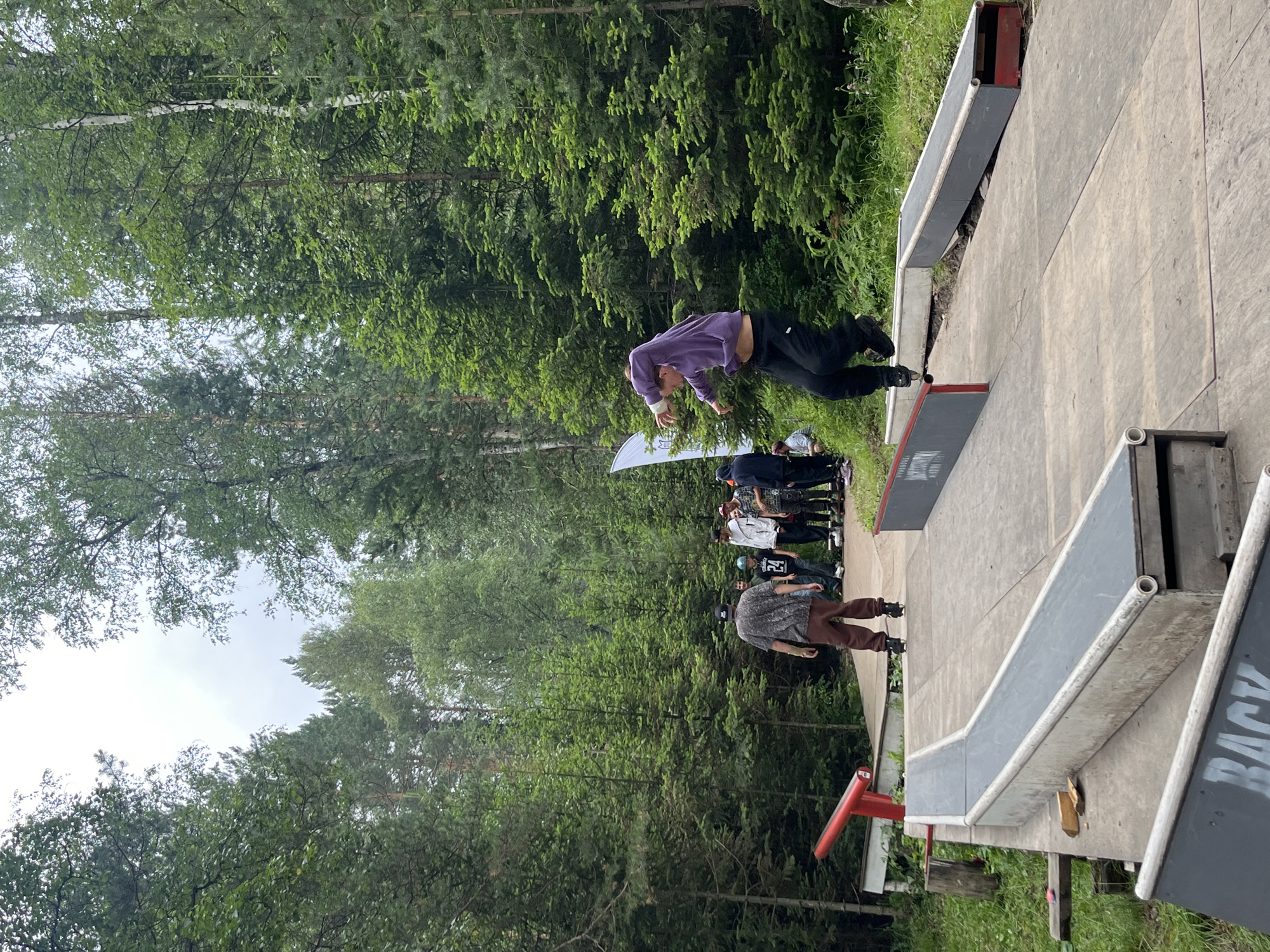 In Divnogorsk, Russia, a group of young people are gathered at a skatepark surrounded by a dense forest. The foreground features a concrete skatepark with various ramps and rails. The central subject is a person in mid-air, performing a trick on rollerblades, wearing a purple hoodie and black pants. Other individuals, dressed in casual attire, are standing and watching the performance. One person in the foreground is also on rollerblades, facing away from the camera. The background is dominated by tall, green coniferous trees, suggesting a natural, outdoor setting. The sky is overcast, indicating a cloudy day. The skatepark itself appears somewhat weathered, with some visible wear and tear on the wooden and concrete surfaces.