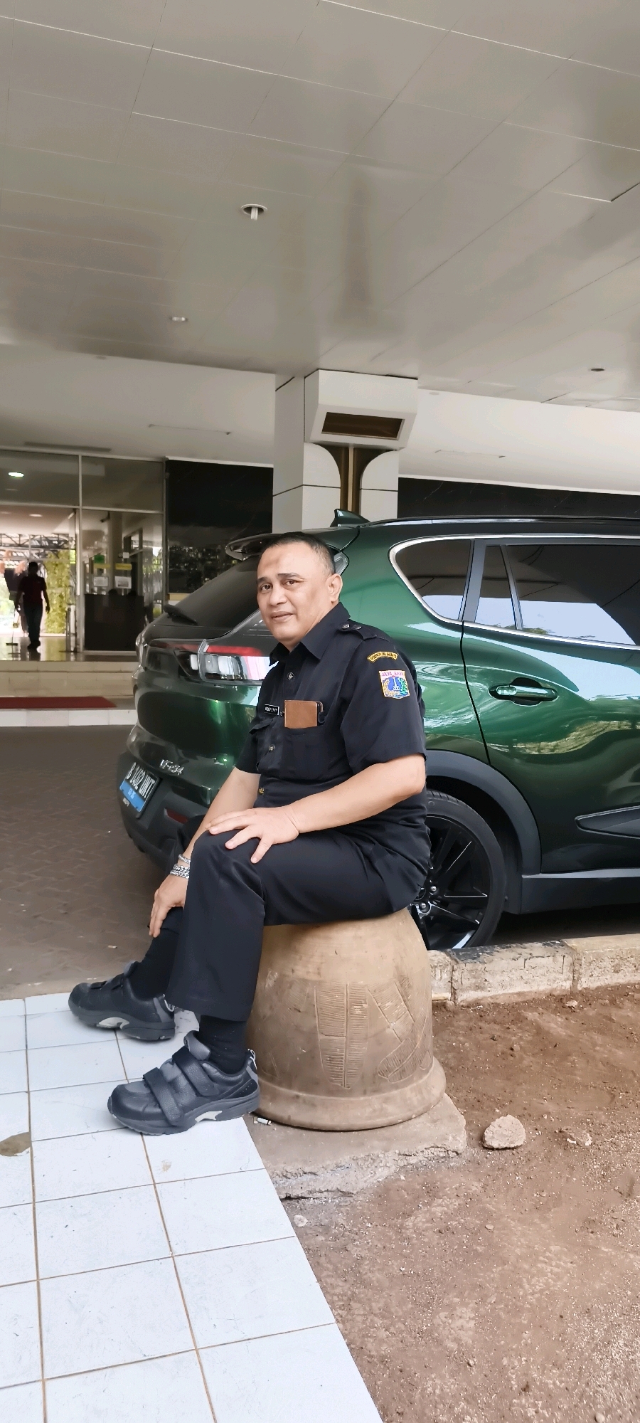 A man in a black uniform is sitting on a large, round, terracotta-colored pot in front of a dark green car. He is wearing a short-sleeved black shirt with patches on the left sleeve and a name tag on the chest, black pants, black socks, and black sneakers with velcro straps. He has short, dark hair and is looking towards the camera. The car, a modern SUV, has tinted windows and a license plate that reads "B 1492 UNT". Behind the car, a building entrance with glass doors is visible. The man is seated in what appears to be an outdoor area with tiled flooring to his left and dirt and gravel to his right. The environment suggests a service area or parking lot, possibly near a hotel or office building in South Jakarta, Indonesia.