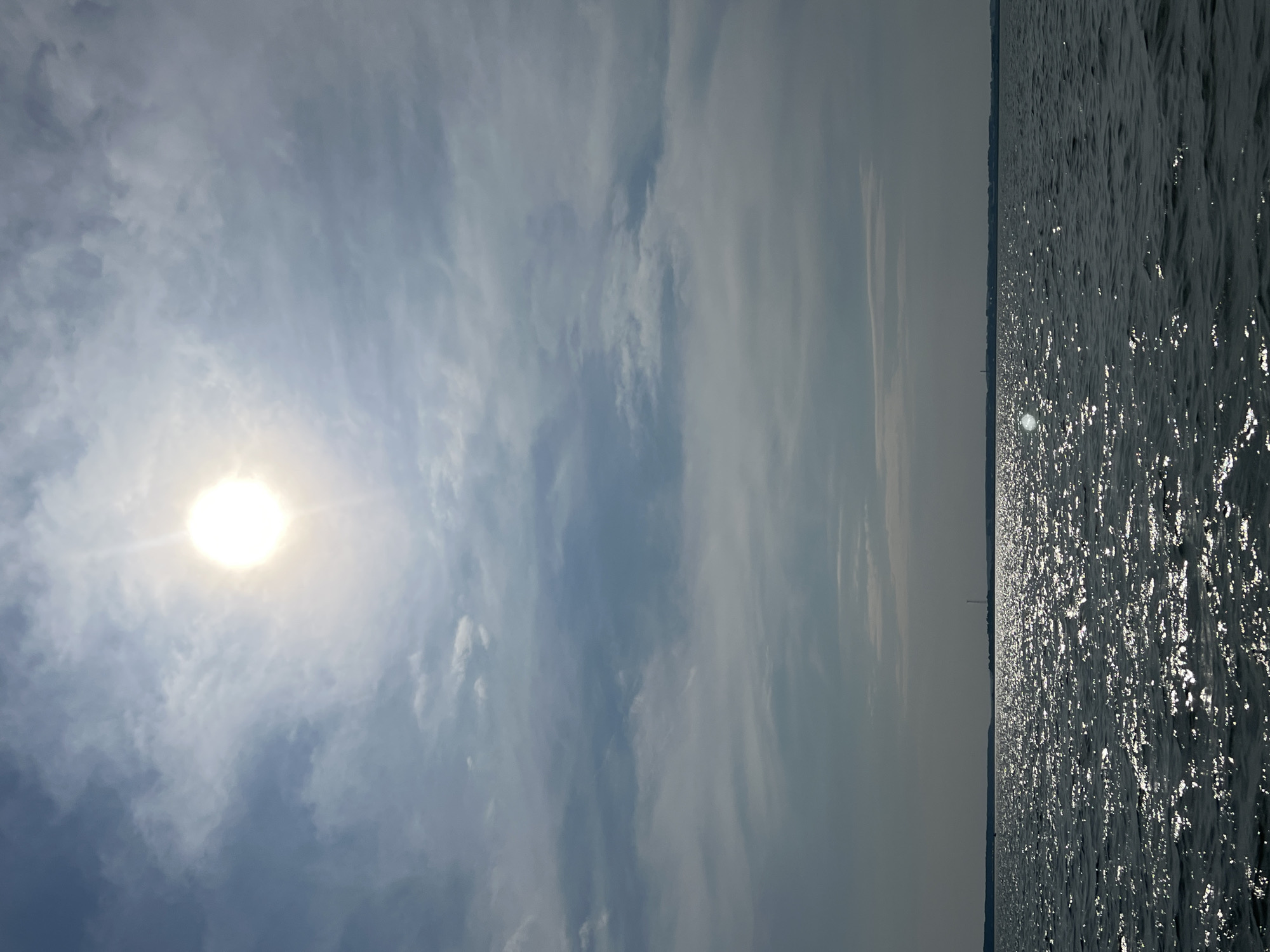 The image depicts a bright sun in a partly cloudy sky above a wide body of water, likely a lake or sea, with a distant shoreline. The sun is positioned in the upper center, casting a bright glare that obscures its details and creates a halo effect. The sky is filled with scattered clouds, some wispy and others more dense, in varying shades of white and gray, contrasting with patches of blue sky.
The water occupies the lower half of the image and is a deep blue-gray, reflecting the sunlight in a shimmering pattern of white sparkles. The surface of the water appears slightly choppy, with small waves creating texture and movement. The horizon line is relatively flat and runs across the middle of the image, separating the sky from the water.
On the distant shoreline, a faint silhouette of land is visible, dotted with what appear to be trees and structures, though they are too indistinct to be clearly identified. There is a tall, slender structure, possibly a tower or chimney, visible on the shoreline. The overall impression is one of a serene, natural landscape under a bright, sunny sky. There are no people or animals visible in the image. The scene does not provide any specific indicators to identify its location within Novopichugovo, Russia.